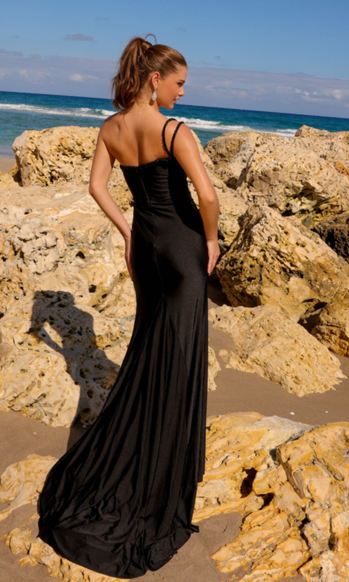 A woman in an Amelia Couture One-Shoulder Prom Dress AC0013 stands on a rocky beach, gazing at the ocean with her back to the camera under a clear sky.