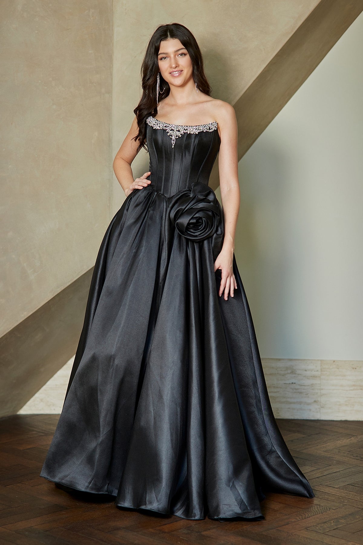 A woman wears a black Corset Prom Ball Gown: Amelia Couture AL1401, featuring a strapless corset bodice, a jeweled neckline, and a large fabric flower accent at the hip.