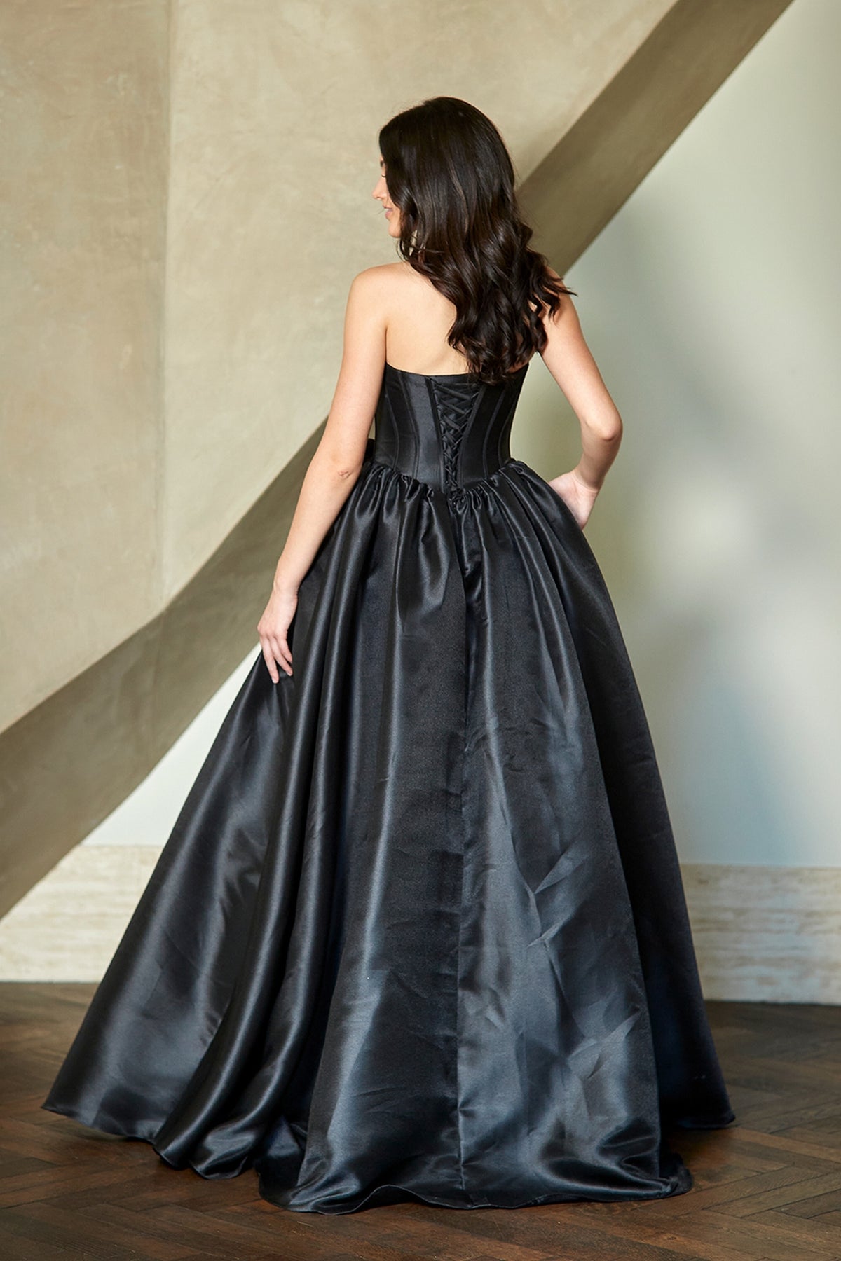 A woman wears the Corset Prom Ball Gown: Amelia Couture AL1401, a strapless black dress with a lace-up back and a full, floor-length skirt.