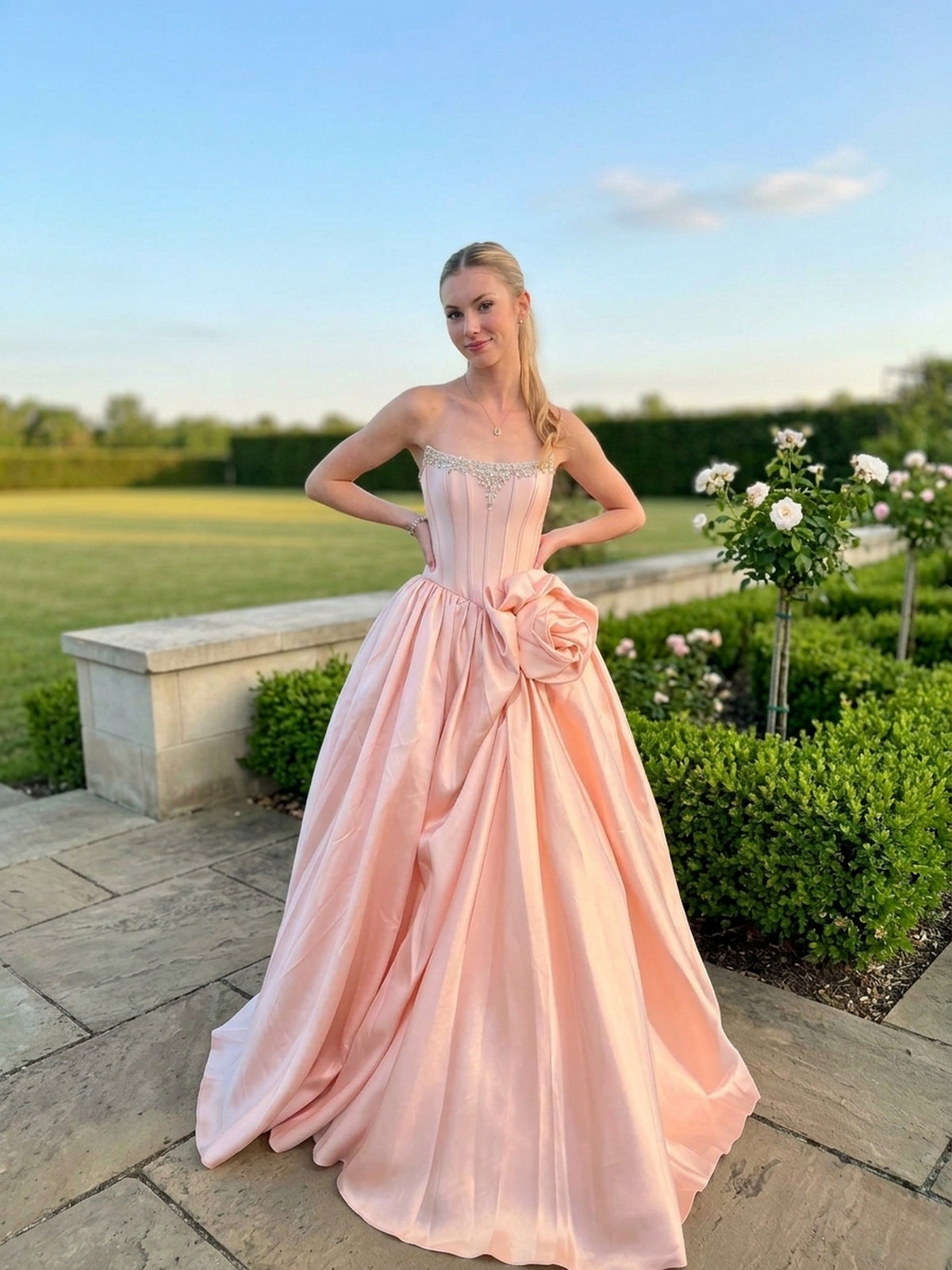 A person wears a rose-colored Amelia Couture AL1401 strapless corset prom ball gown with a beaded bodice and a large floral accent at the waist.
