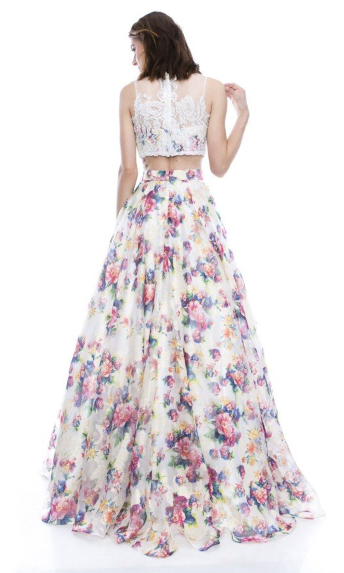 A woman faces away from the camera wearing the Two-Piece Floral-Print Long Prom Dress B6803, featuring a sleeveless white lace crop top and a vibrant, floral-print skirt.