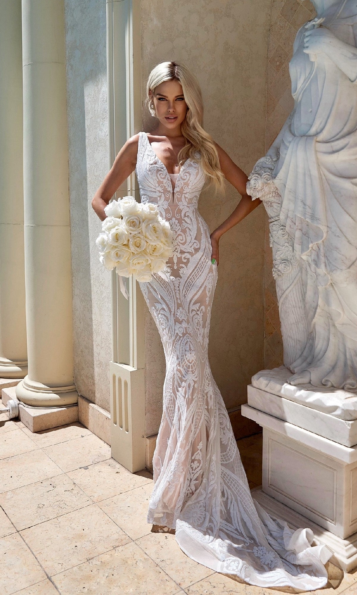 A woman in the Tina Holly BA109 White Lace Mermaid Wedding Gown holds a bouquet of white roses while standing next to a white statue and columns.