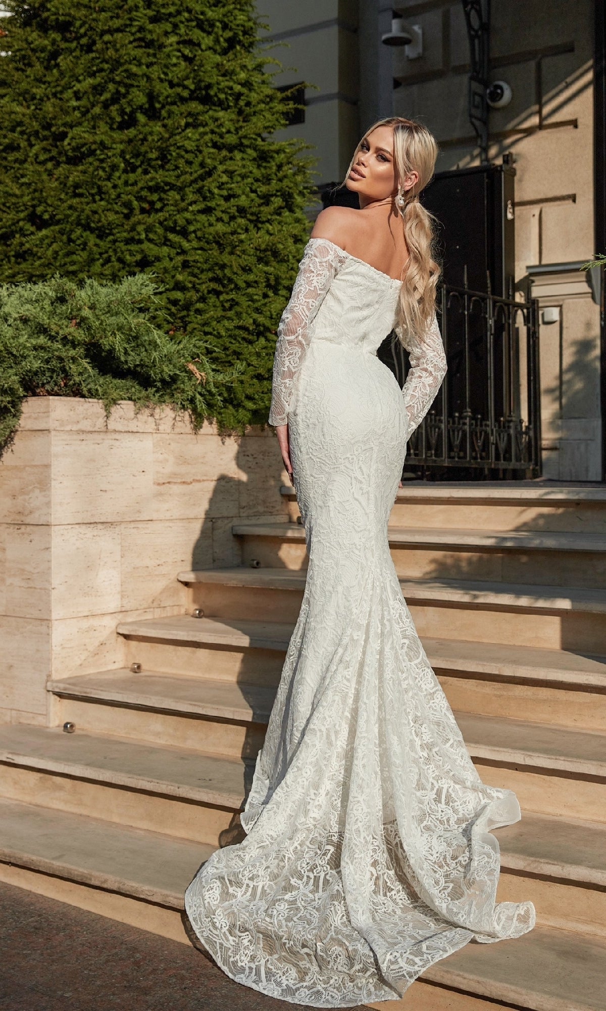 A woman stands on outdoor steps, looking over her shoulder in the stunning Long Lace White Formal Gown: Tina Holly BB007, featuring an off-the-shoulder design and long lace sleeves.
