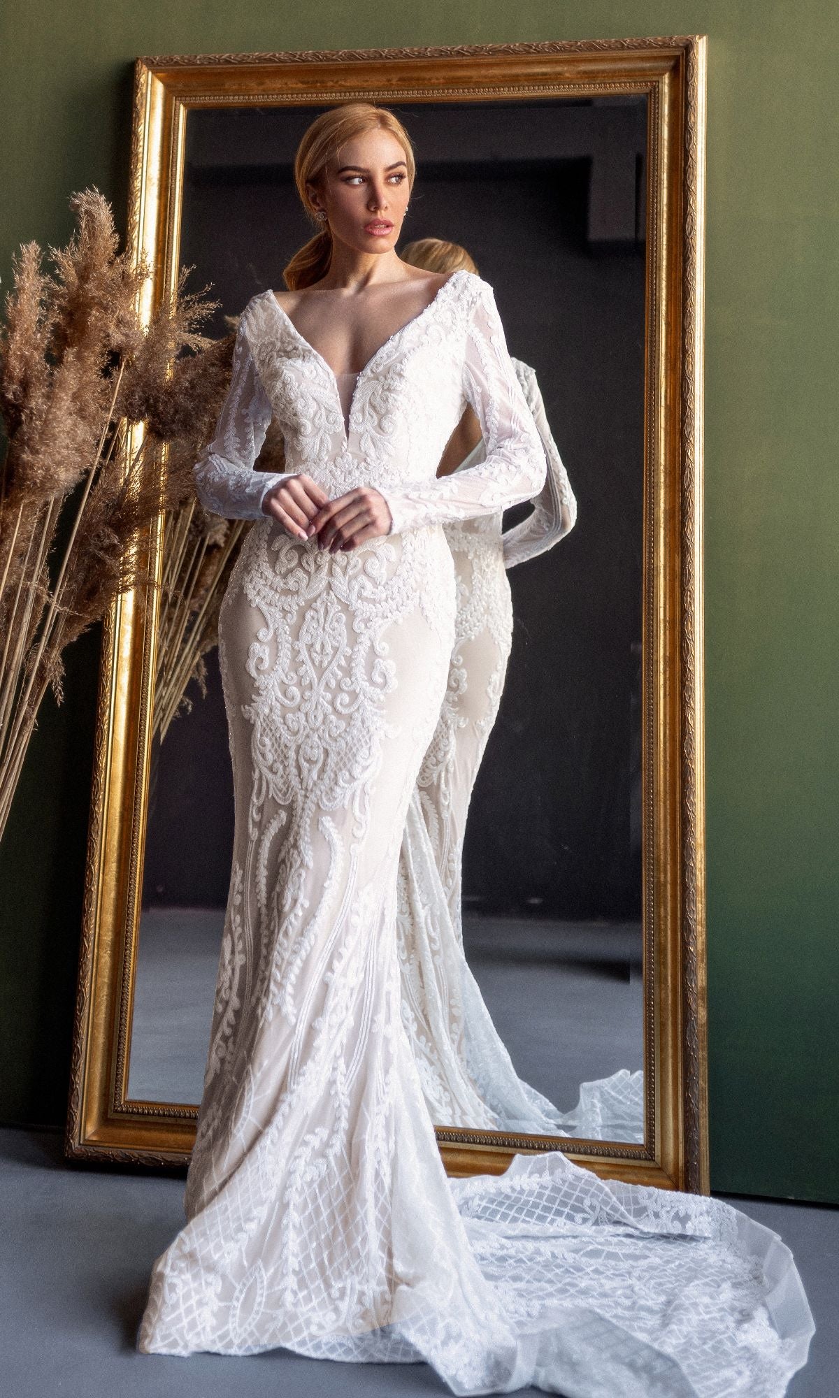 A woman wearing the Tina Holly BB021 long sleeve white bridal gown with a mermaid skirt stands before a large ornate mirror, dried plants on the side.