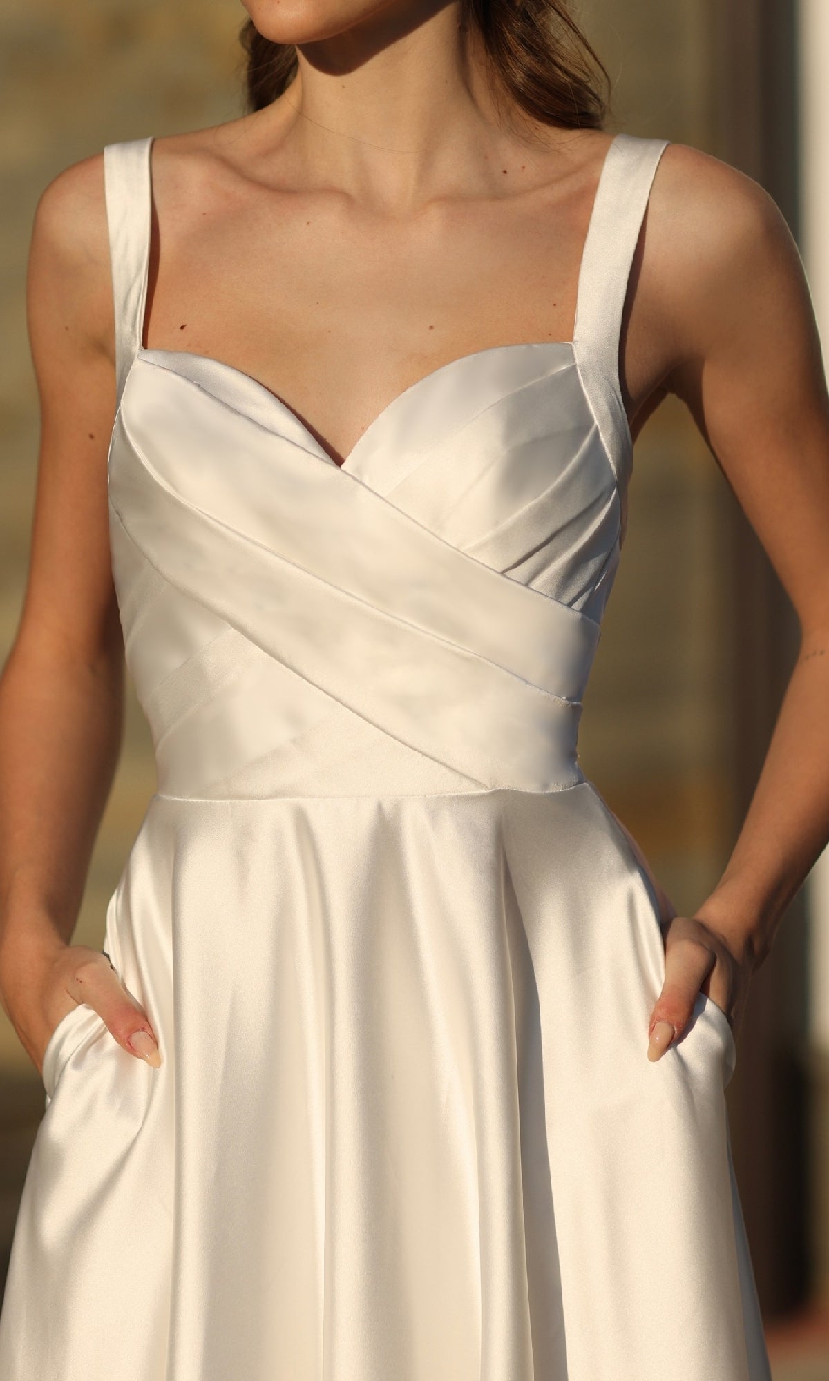 A woman stands with her hands in the pockets of the Off-White Satin Prom Ball Gown Tina Holly BB210W, a sleeveless formal evening dress with a fitted, pleated bodice and thin straps.