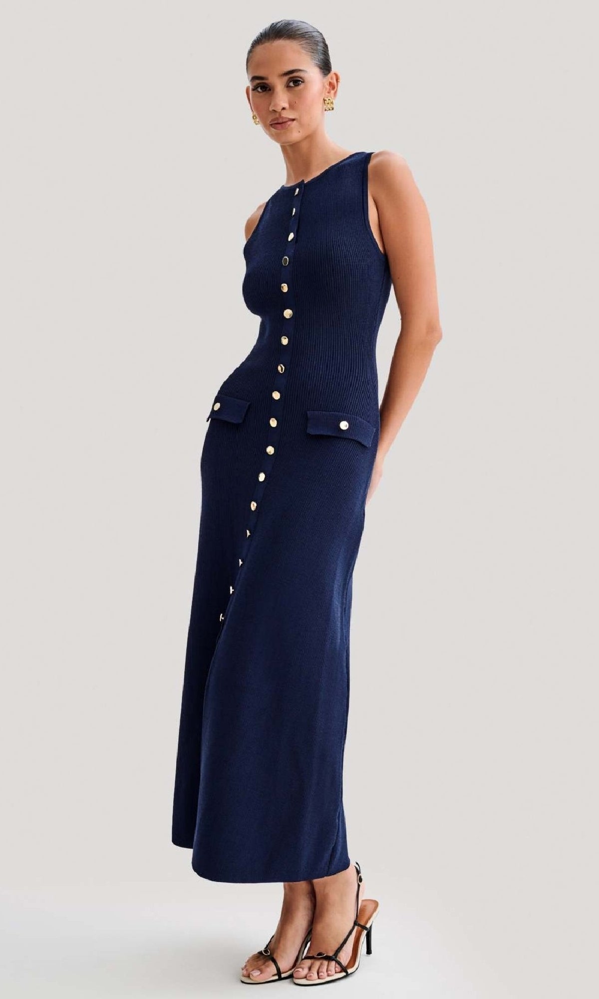 A woman stands in sandals against a plain light background, wearing the Sleeveless Casual Button-Down Maxi Dress in navy with gold buttons and two front pockets.