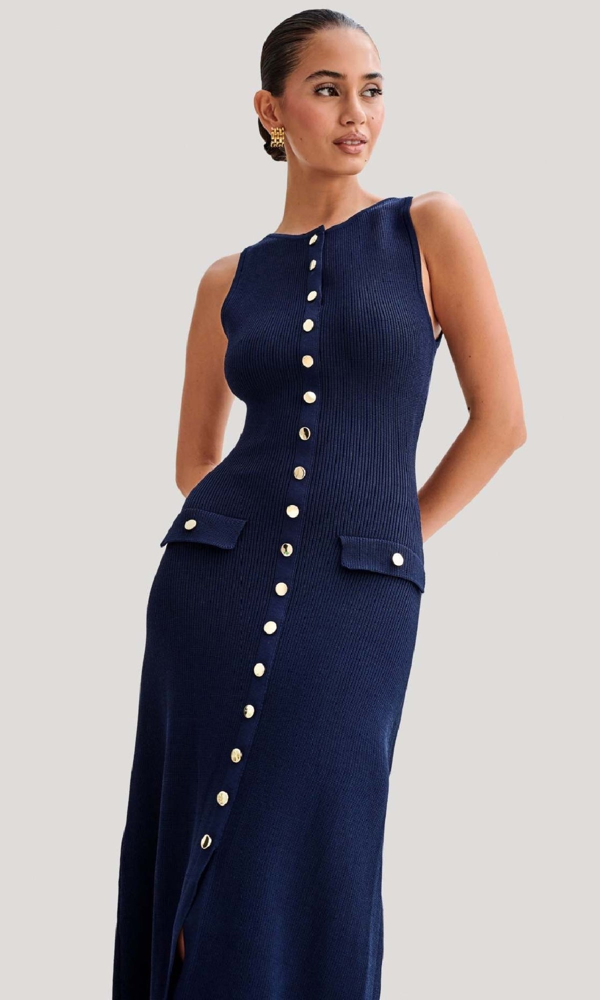 A woman stands against a plain background wearing the Sleeveless Casual Button-Down Maxi Dress, featuring a navy blue ribbed design, gold front buttons, and two hip flap pockets.