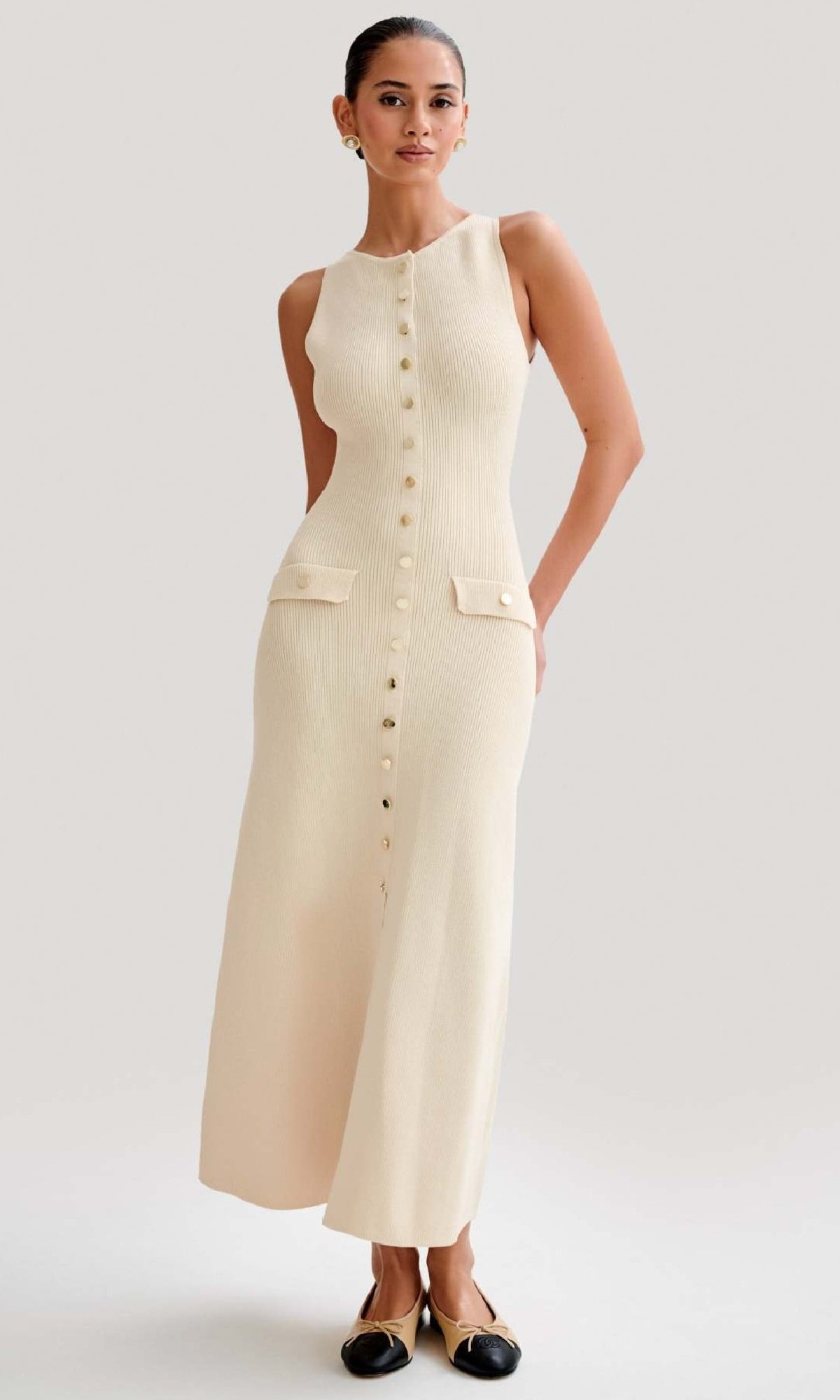 A woman wears the Sleeveless Casual Button-Down Maxi Dress in cream, featuring front buttons and two pockets, paired with black-toed beige flats.