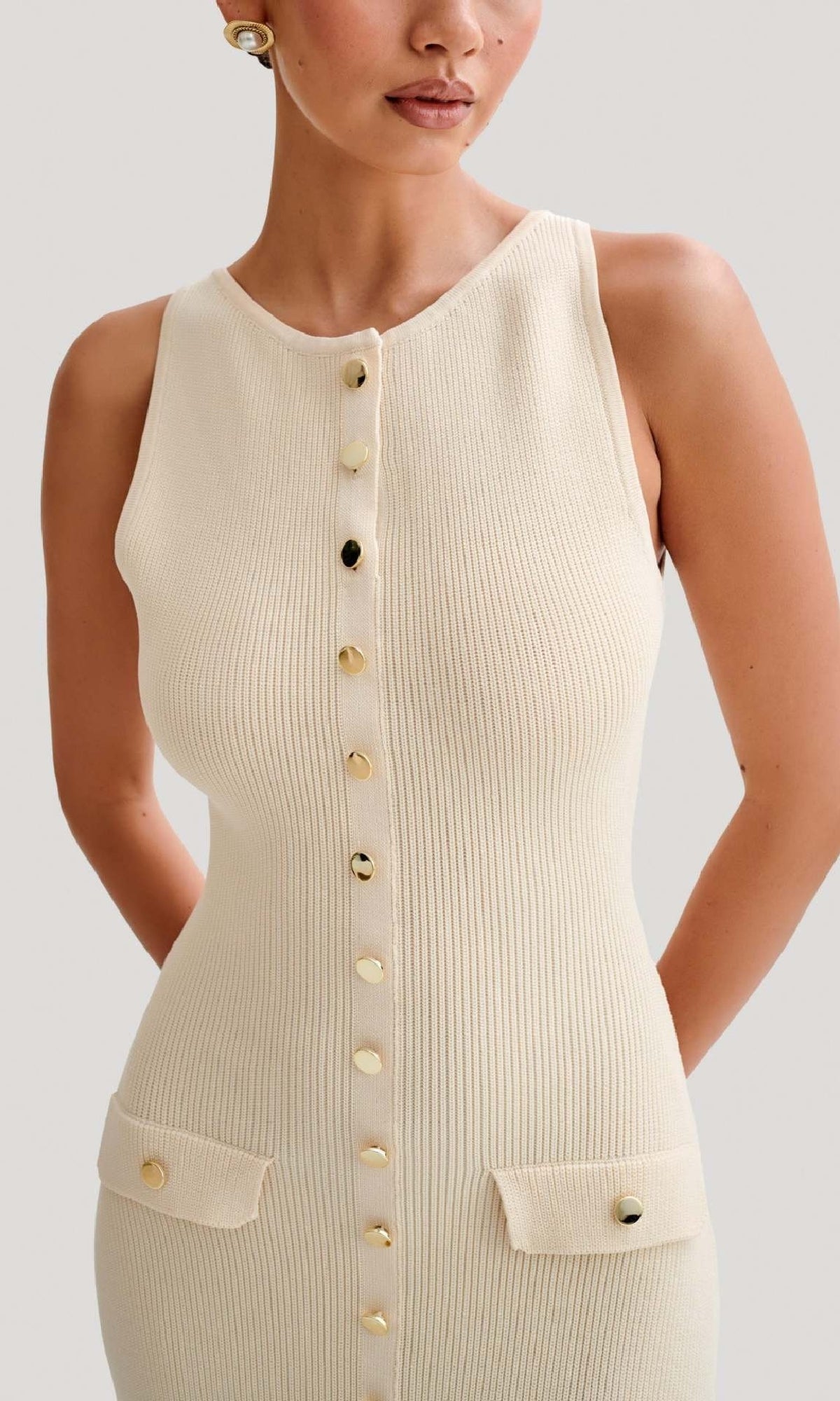 A woman wears the Sleeveless Casual Button-Down Maxi Dress in cream, featuring ribbed fabric, gold front buttons, and two flap pockets, shown from mid-face to thigh.