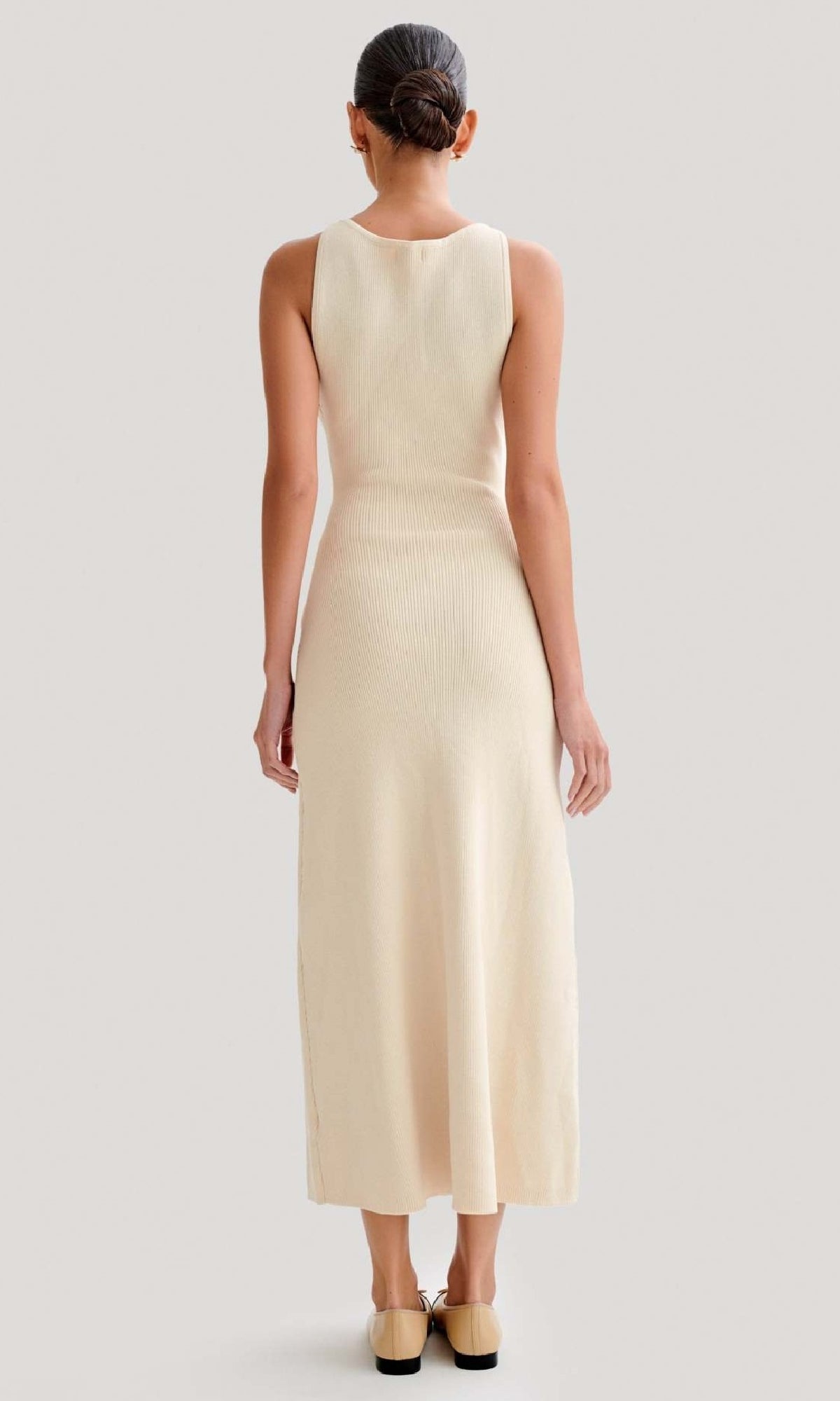 A woman stands facing away, wearing the Sleeveless Casual Button-Down Maxi Dress in a cream shade with flat shoes, her hair neatly tied back in a bun against a plain background.