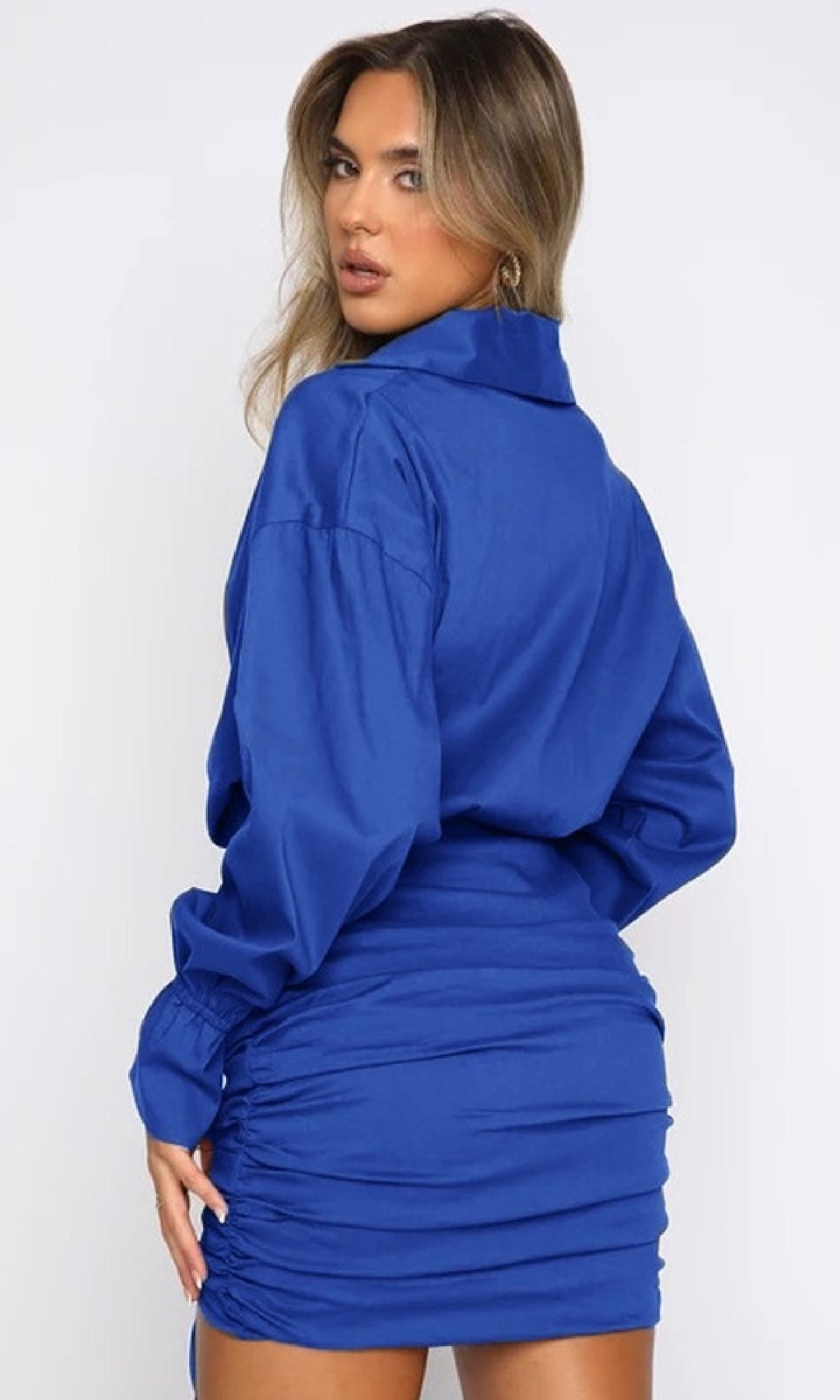A woman stands with her back turned, looking over her shoulder, wearing a Short Casual Royal Blue Shirt Dress with sleeves and ruched detailing on the skirt.