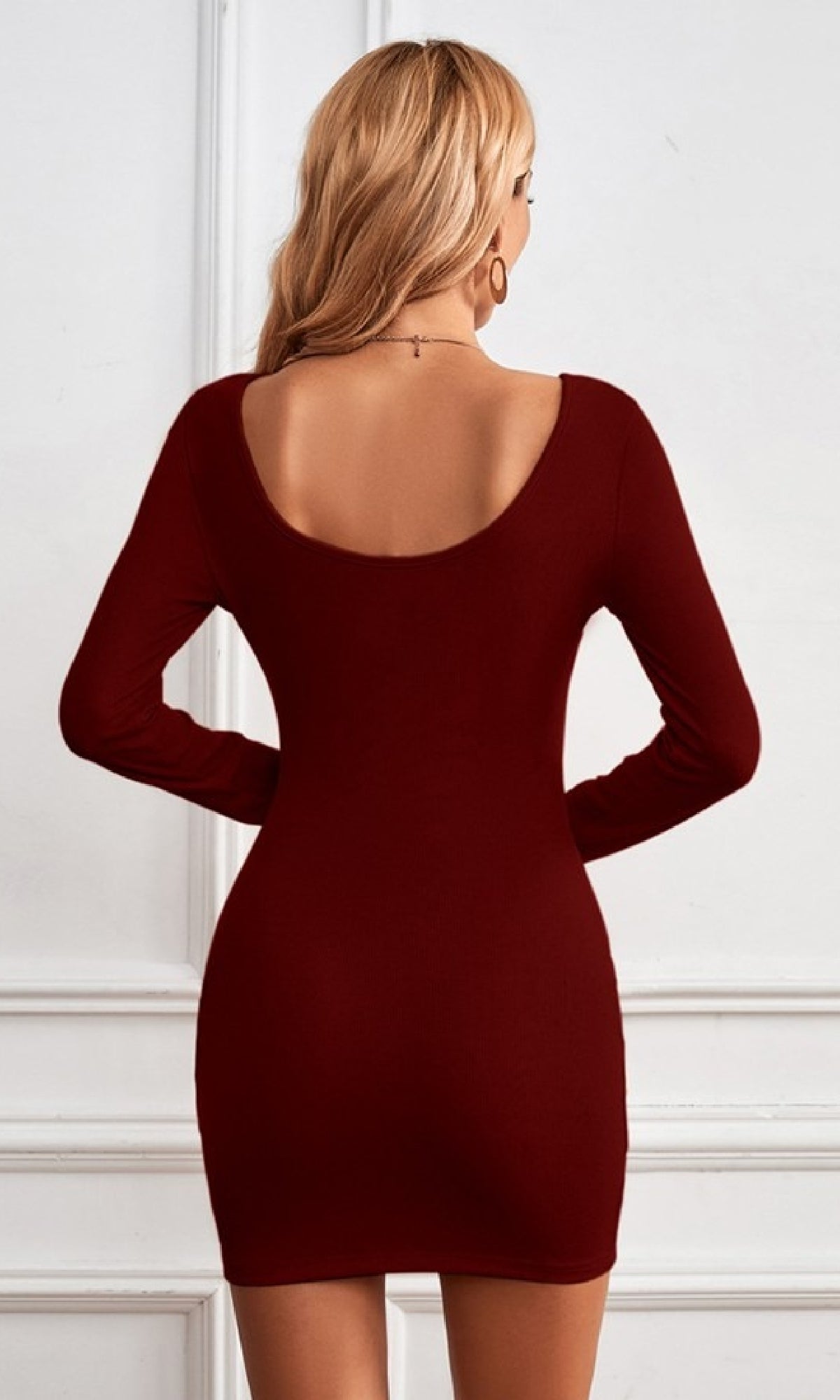 A woman with long blonde hair, seen from the back against a white paneled wall, wears the Burgundy Red Long-Sleeve Mini Party Dress.