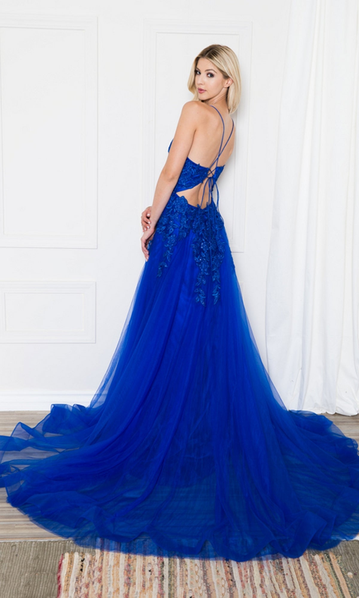 Woman modeling the Long Tulle Prom Ball Gown with Train BZ014 in royal blue, featuring embroidered floral details and an open back, poses by a white wall and curtain.