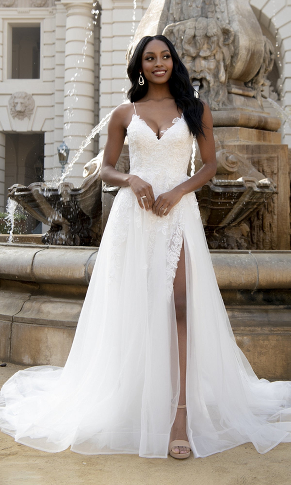 A woman wearing the Long Tulle Prom Ball Gown with Train BZ014, featuring a white sleeveless design and delicate floral embroidery, stands smiling in front of a stone fountain.
