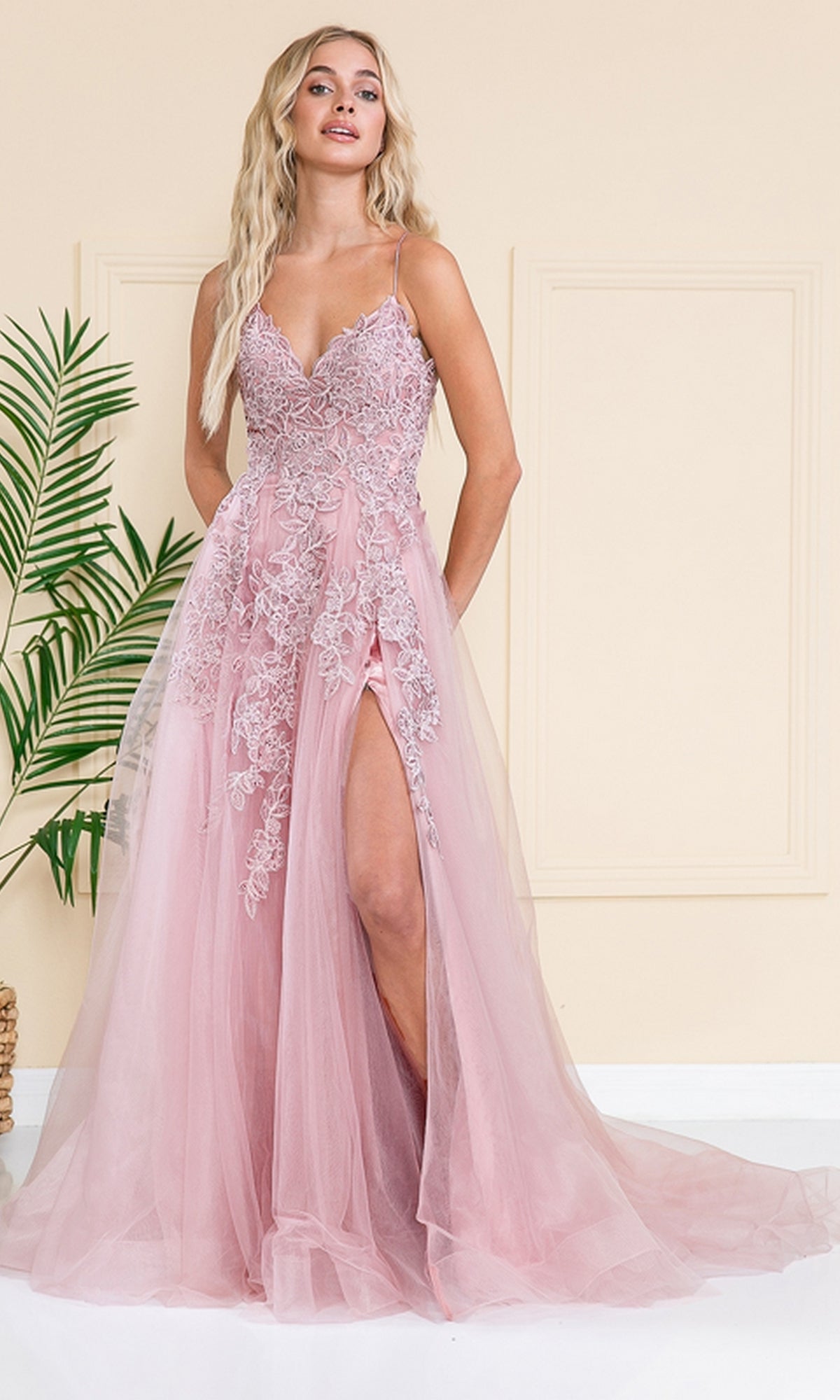Woman wearing the Long Tulle Prom Ball Gown with Train BZ014, featuring a pink hue, thigh-high slit, and embroidered floral details, standing indoors by a beige wall and green plant.