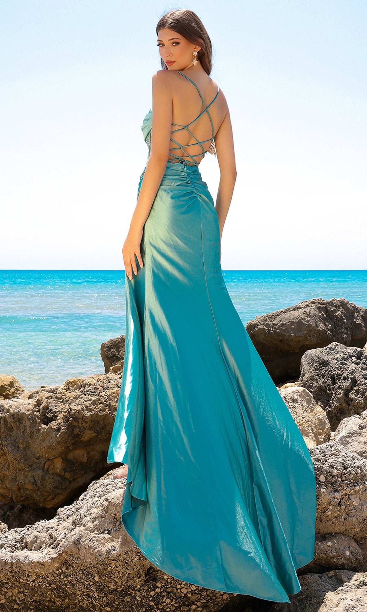 A woman stands on seaside rocks in the Sheer-Corset Cowl-Neck Long Prom Dress BZ020, its teal, strappy back and flowing cowl-neck style elegantly highlighted against the blue sky and ocean backdrop.