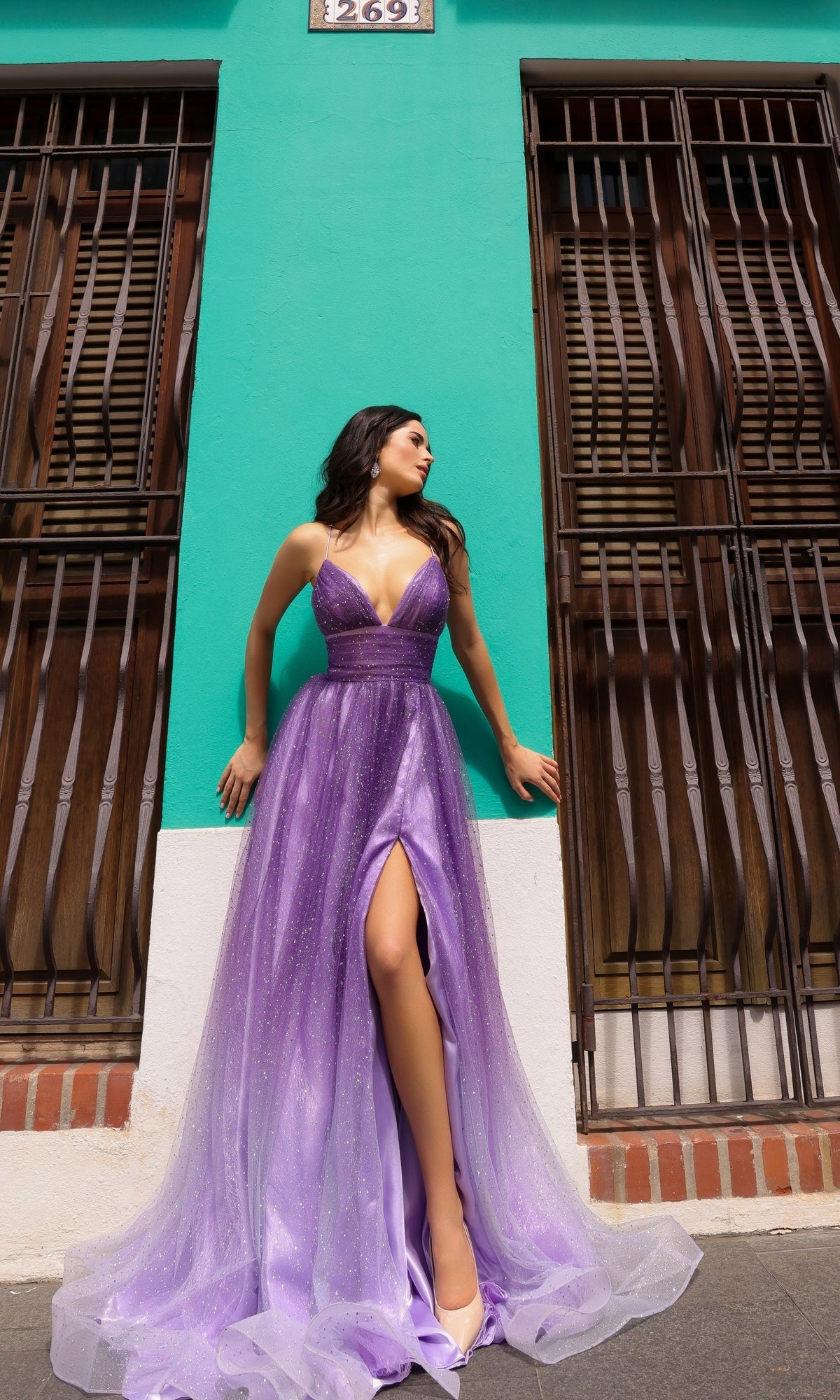 A woman in the Lace-Up Ombre Purple Long A-Line Prom Dress C1251 with a thigh-high slit stands between two barred windows against a turquoise wall.
