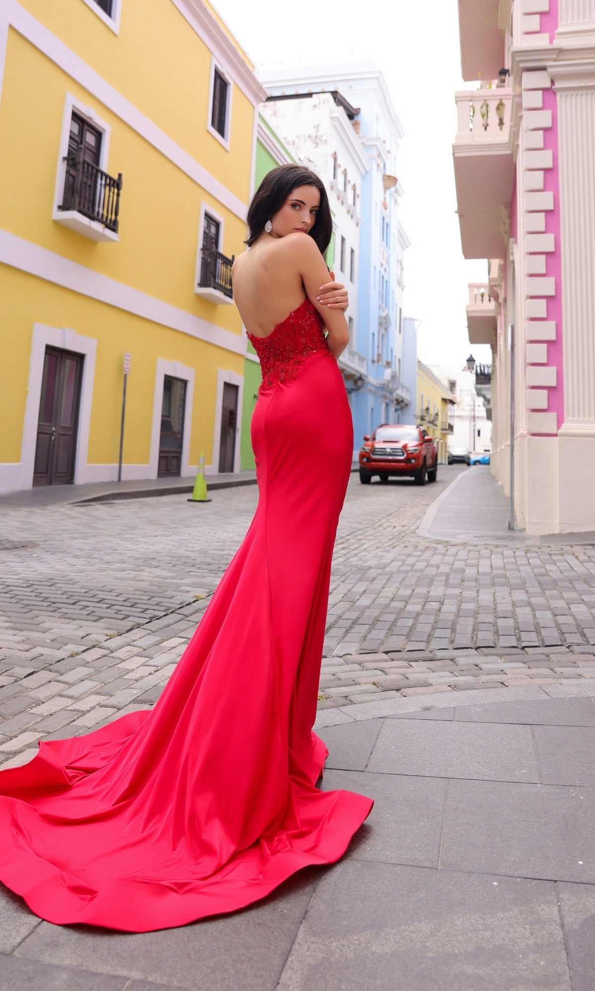 A woman in the Strapless Sweetheart Corset Long Prom Dress C1345 with an embellished bodice stands on a cobblestone street lined with colorful buildings, looking over her shoulder.