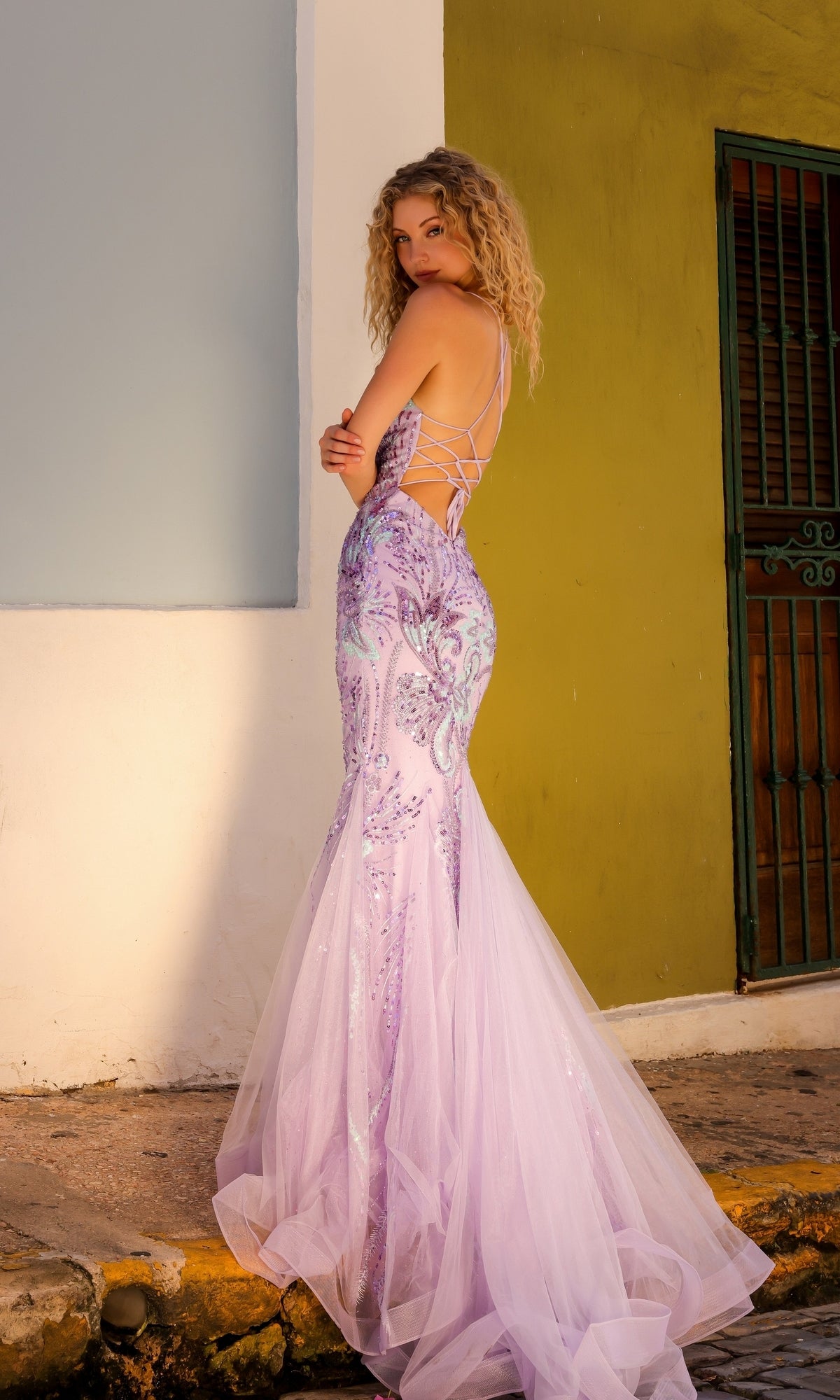A woman with curly blonde hair wears the Nox Anabel Tight Sequin Mermaid Prom Dress C1416 in lavender, featuring a backless design, as she stands on a sidewalk near a green door and pale blue and white wall.