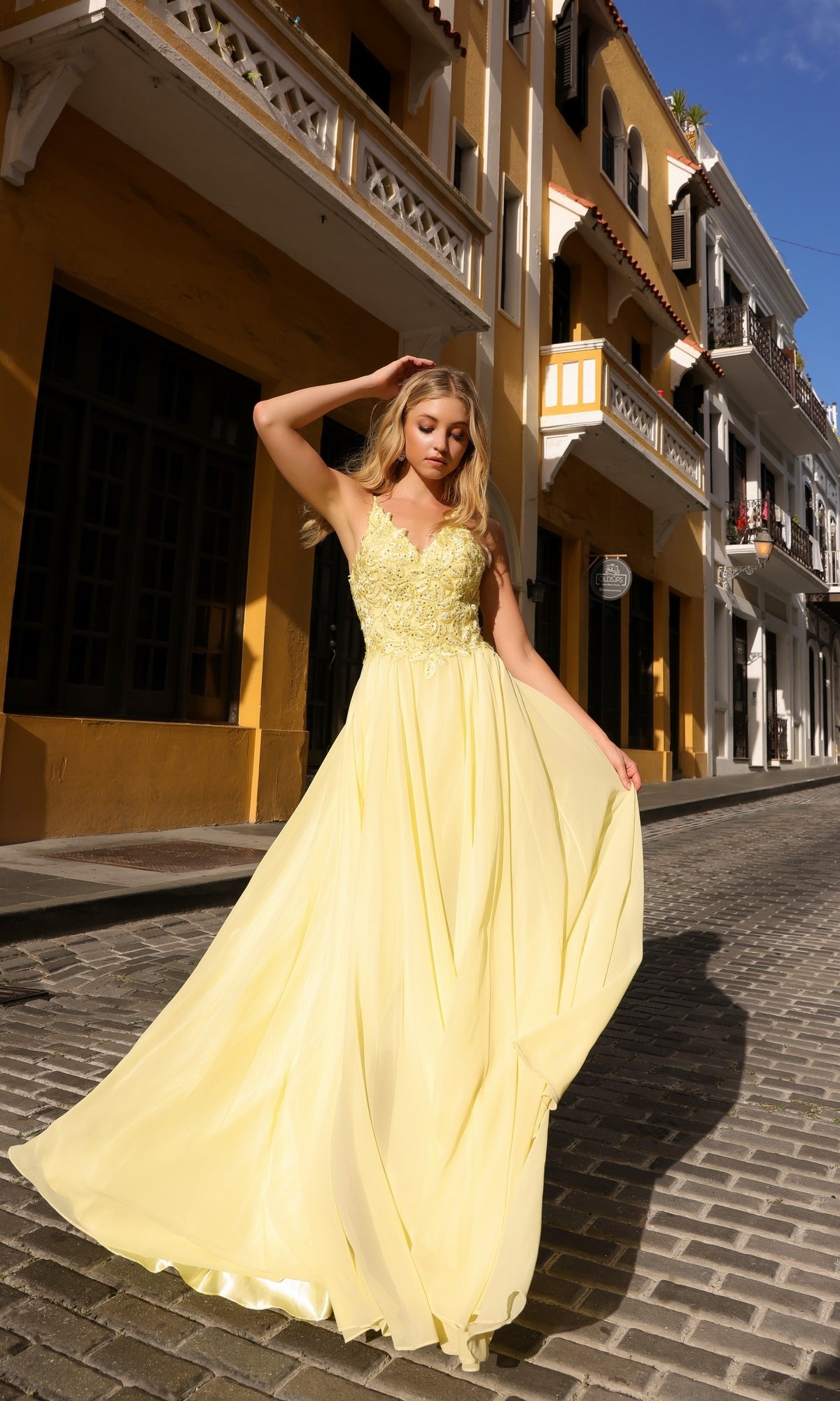 A woman wearing the Long Pastel A-Line Prom Dress C1462, featuring a flowing skirt and embroidered-lace bodice, stands on a cobblestone street bordered by yellow and white buildings, holding her gown and looking down.