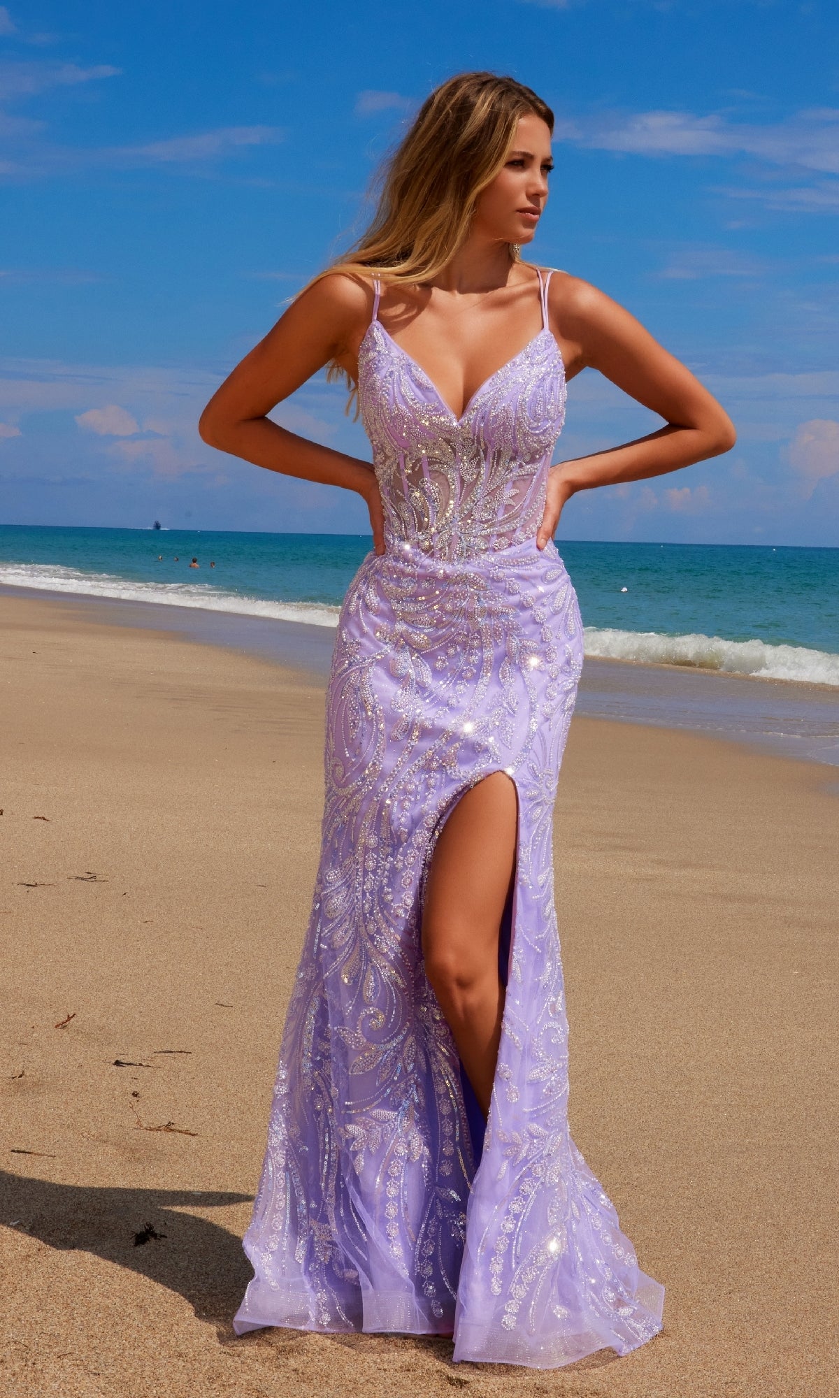 A woman in the Lace-Up Long Beaded Prom Dress: Nox Anabel C1612, featuring a corset back and thigh-high slit, stands on a sandy beach near the ocean under a sunny sky.