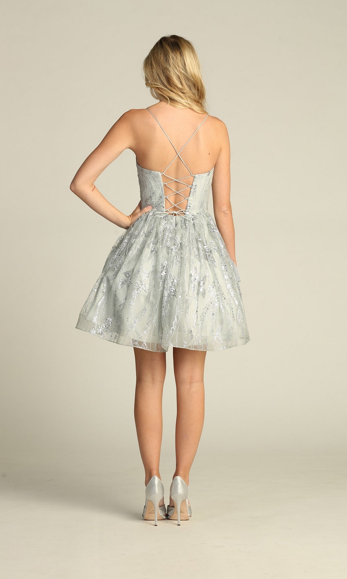 A woman faces away wearing the Lace-Up Glitter-Print Short Homecoming Dress C317, a light gray A-line party dress with silver embellishments, lace-up back, and spaghetti straps. She pairs it with silver heels.