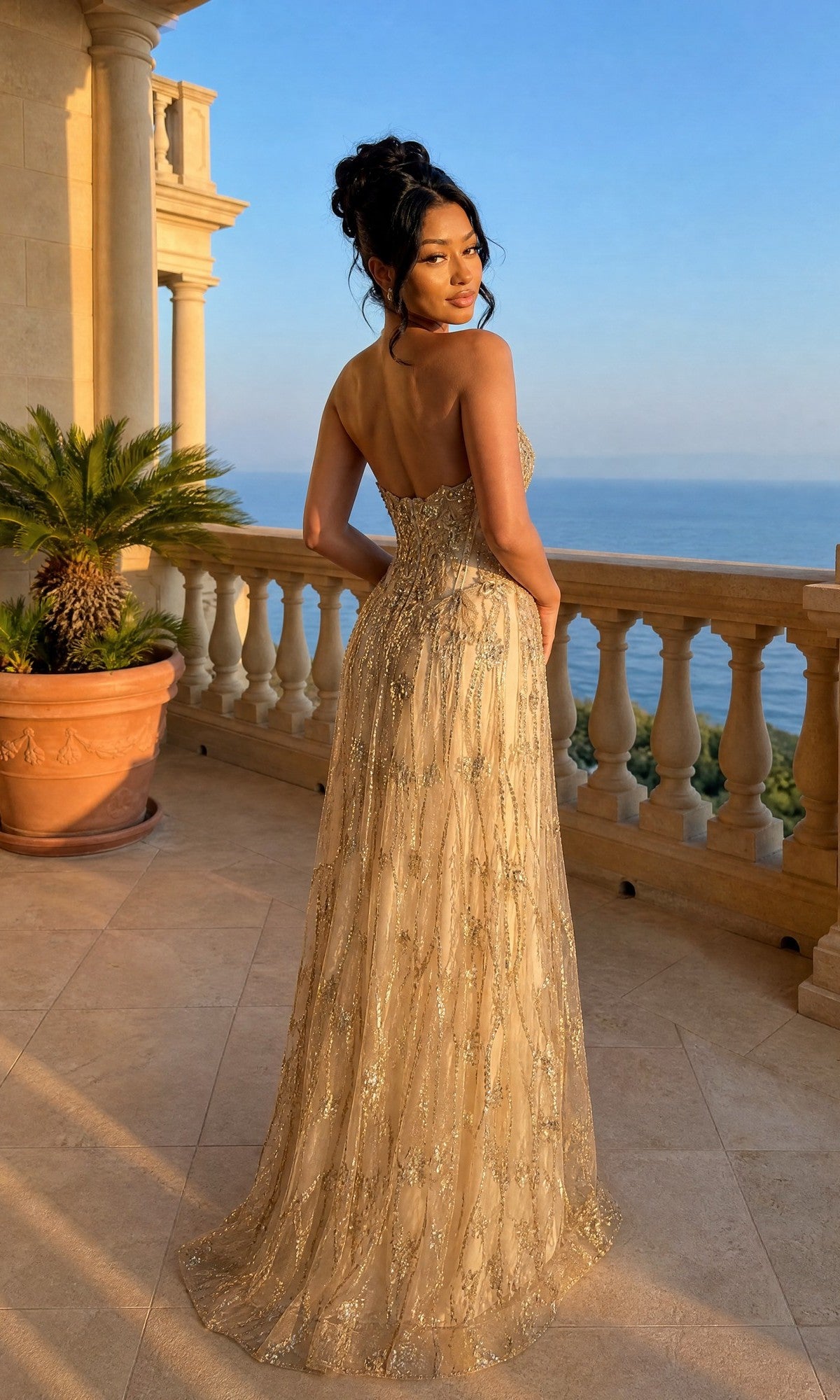 A woman in the Long Prom Dress: Chicas C329—featuring a strapless sweetheart neckline, corset bodice, and gold floral lace applique—stands on a balcony at sunset, overlooking the sea with palm plants and classical architecture.