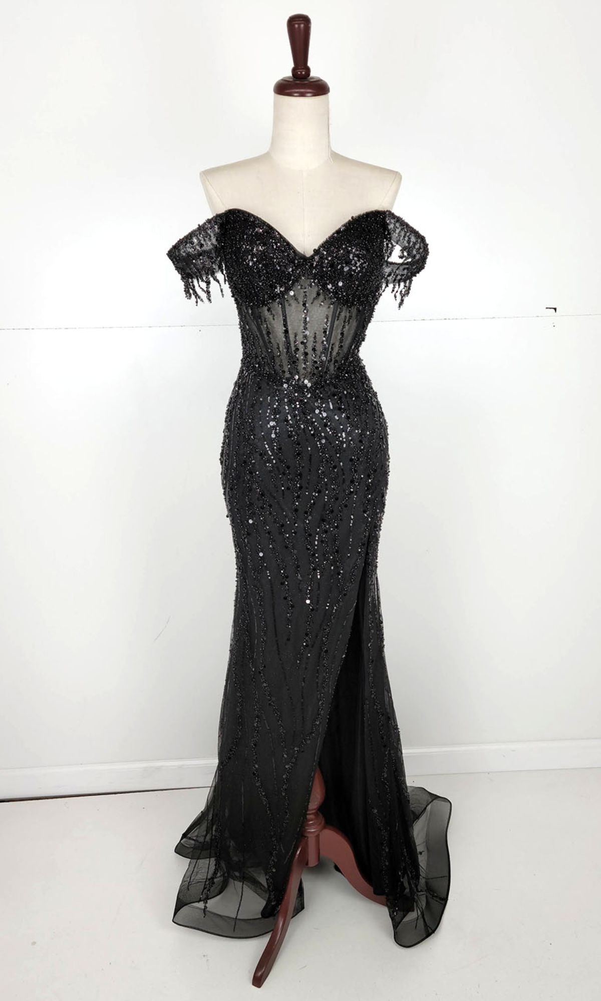 The Off-Shoulder Long Sequin Prom Dress: Chicas C331 features black sequins, sheer accents, and a front slit. Displayed on a dress form with a plain background, it’s perfect for anyone seeking an elegant evening gown.