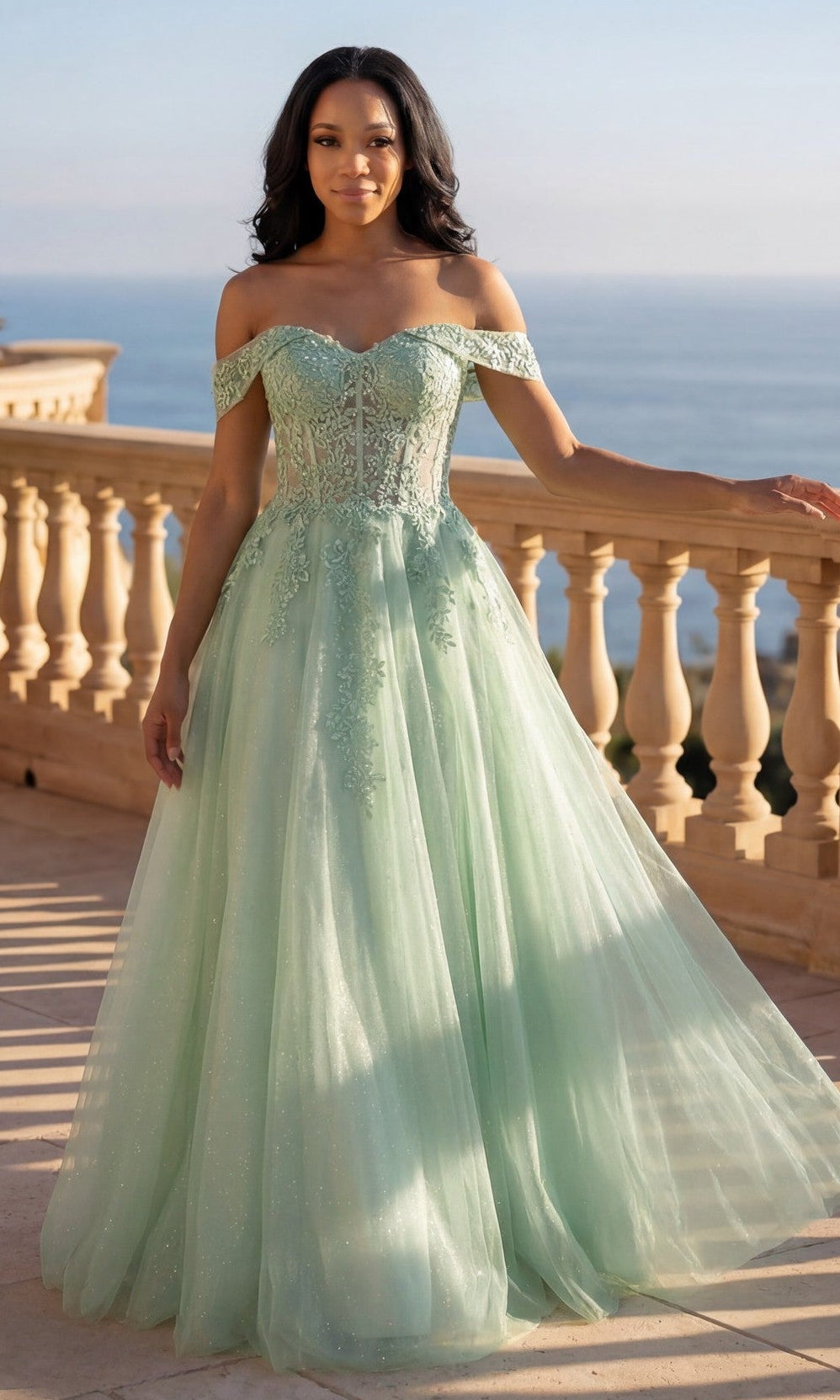 A woman in a sage Long Prom Dress: Chicas C336 with floral lace embroidery stands on a sunlit terrace, the sea view behind her.