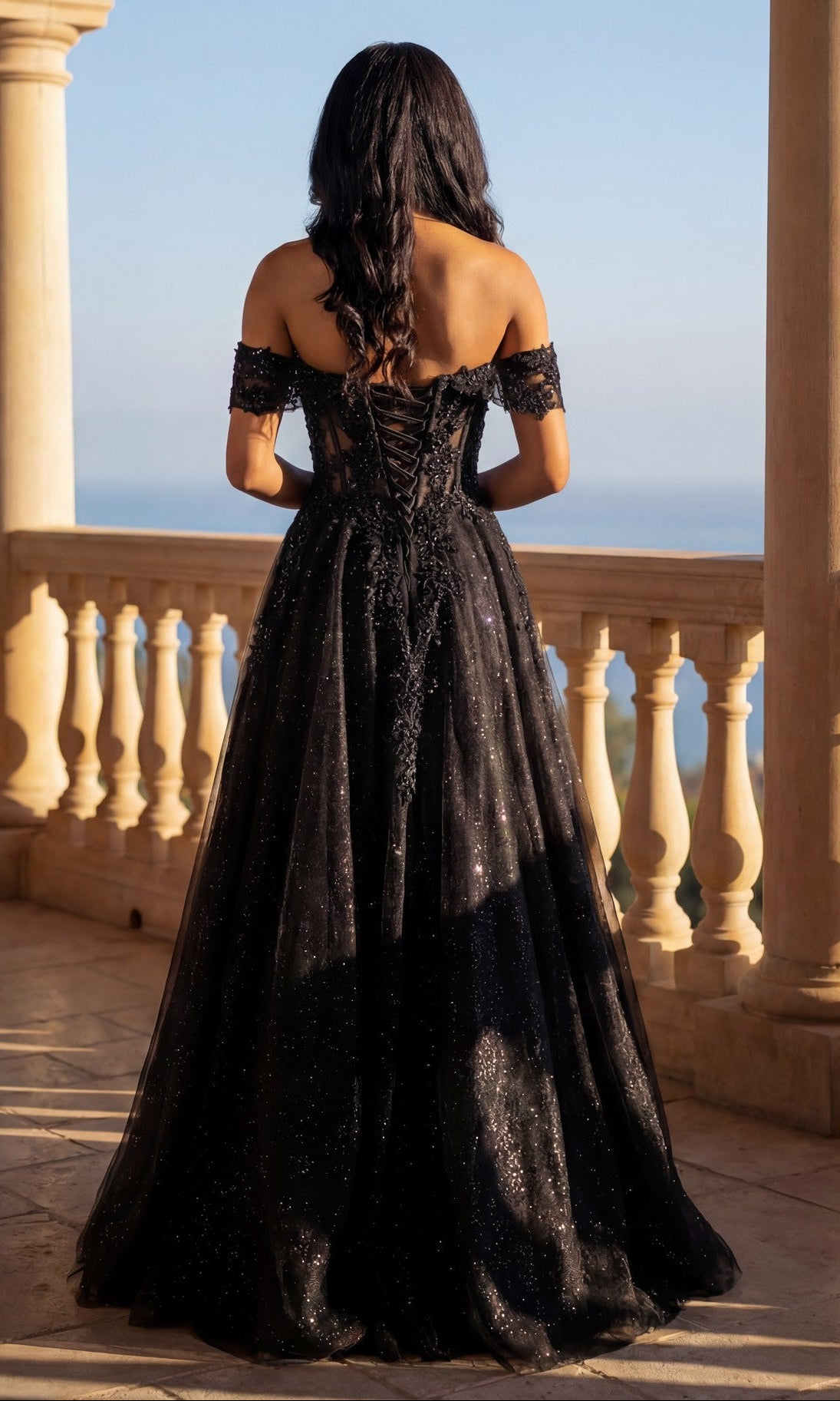 Wearing the Long Prom Dress: Chicas C336, a woman in a black glitter tulle gown with an adjustable lace-up back stands on a stone balcony, gazing at the scenic, sunlit view.