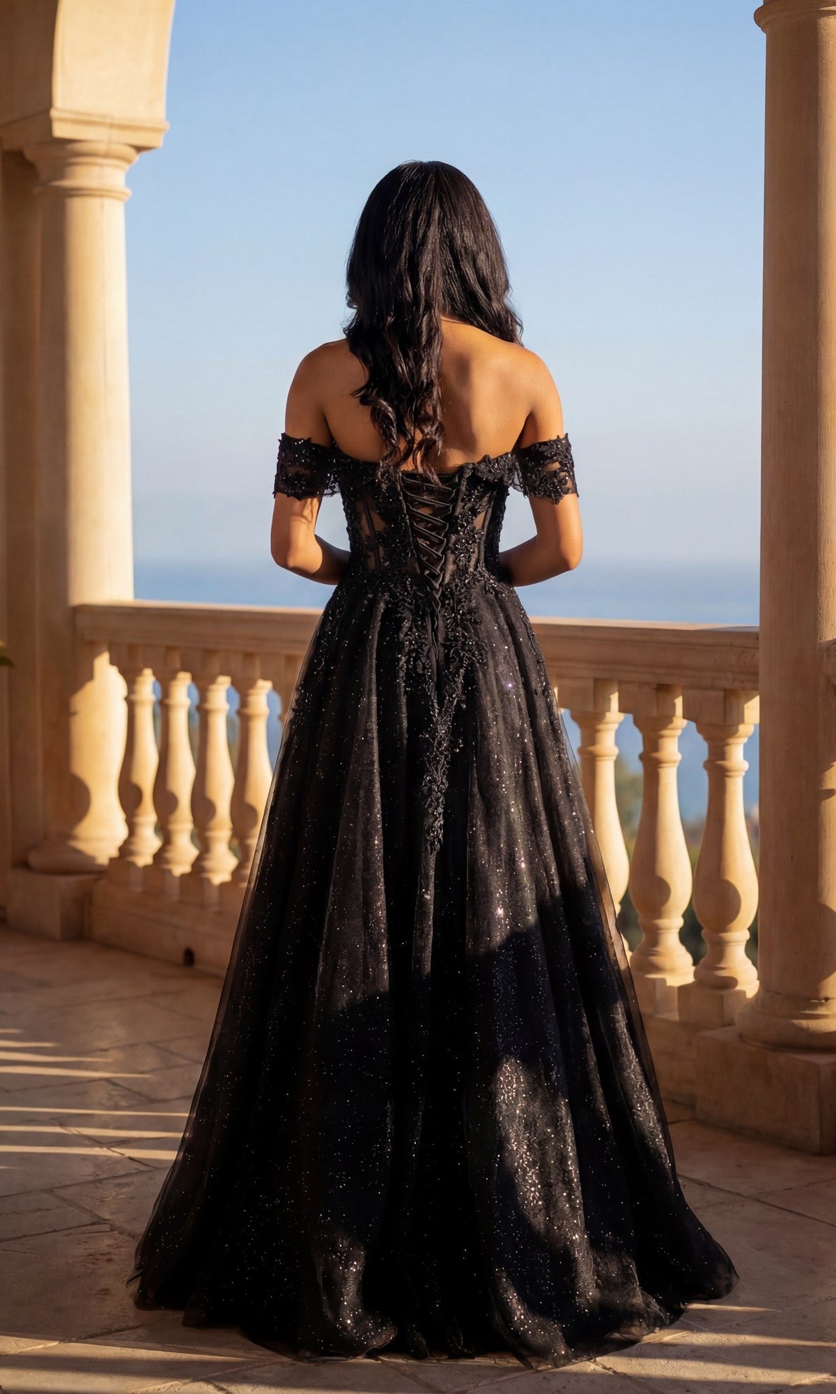Wearing the Long Prom Dress: Chicas C336, a woman in a black glitter tulle gown with an adjustable lace-up back stands on a stone balcony, gazing at the scenic, sunlit view.