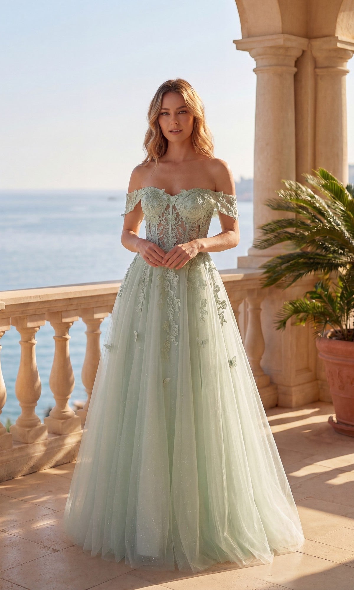 A woman in the Chicas C350 Long Prom Dress—sage, off-the-shoulder glitter tulle with floral accents and a corset bodice—stands on a stone terrace by the sea, beside a potted plant.