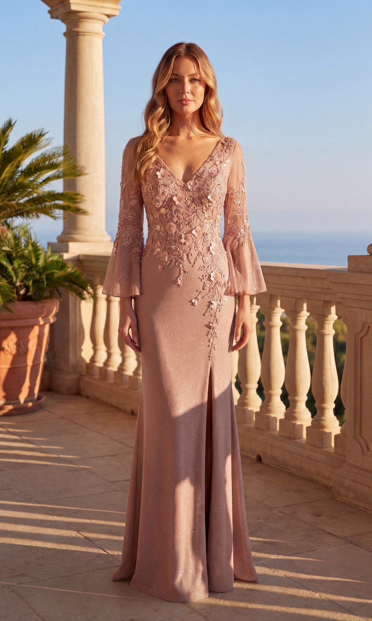 A woman in the Long Formal Dress: Chicas C357 in rose gold, featuring lace details and flared sleeves, stands on a sunlit terrace with columns overlooking the sea.