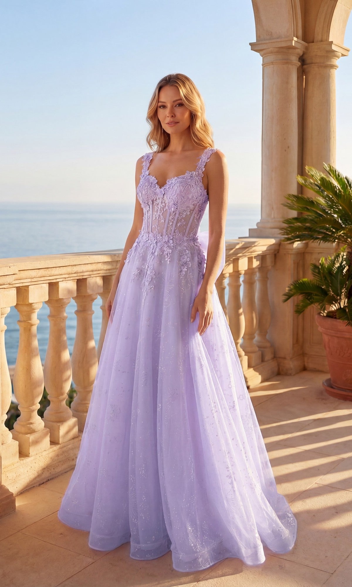 A woman in a lilac Chicas C379 long prom dress with a corset bodice stands on a sunlit balcony overlooking the sea, surrounded by stone columns and a potted palm.