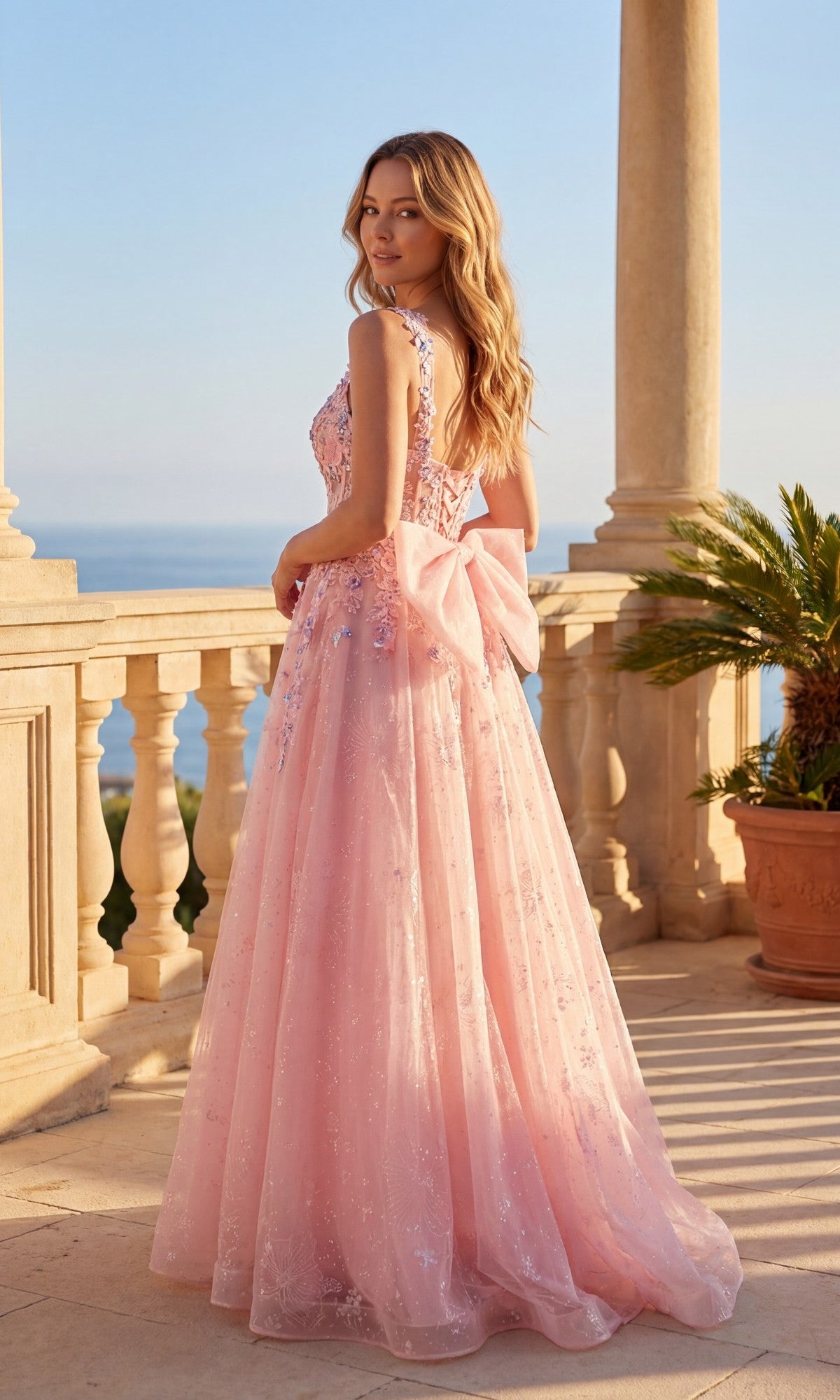 A woman in the Chicas C379 Long Prom Dress, a light pink A-line gown with a large back bow and floral sequin lace embroidery, stands on a stone terrace by the sea with a potted palm nearby.