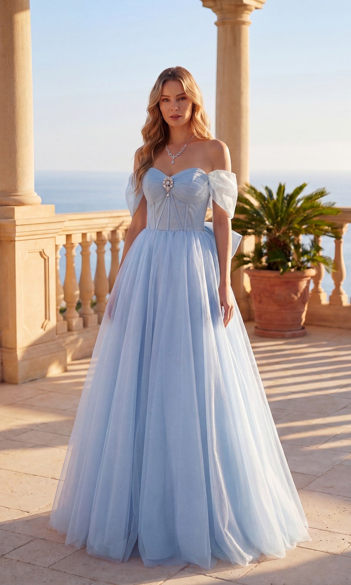 A woman stands on a stone terrace wearing the Long Prom Dress: Chicas C386 in sky blue, featuring an off-the-shoulder fitted bodice and a flowing glitter tulle skirt. A potted plant and the ocean are visible in the background.