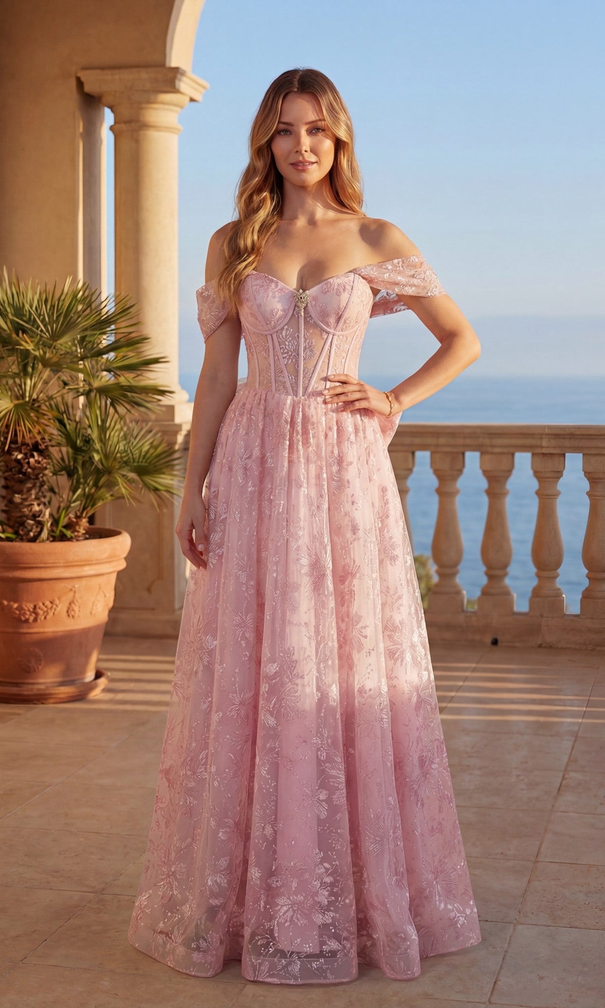 A woman stands on a terrace overlooking the sea, wearing the Long Prom Dress: Chicas C391 in blush, featuring an off-the-shoulder lace design with a sheer corset bodice and a removable back bow.