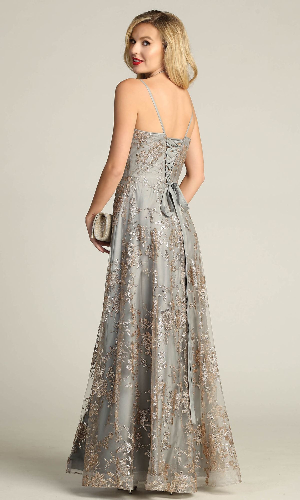 Woman in the Chicas C6534 Glitter-Print Long Gray Prom Dress, featuring a lace overlay and lace-up back, holds a clutch and looks over her shoulder.