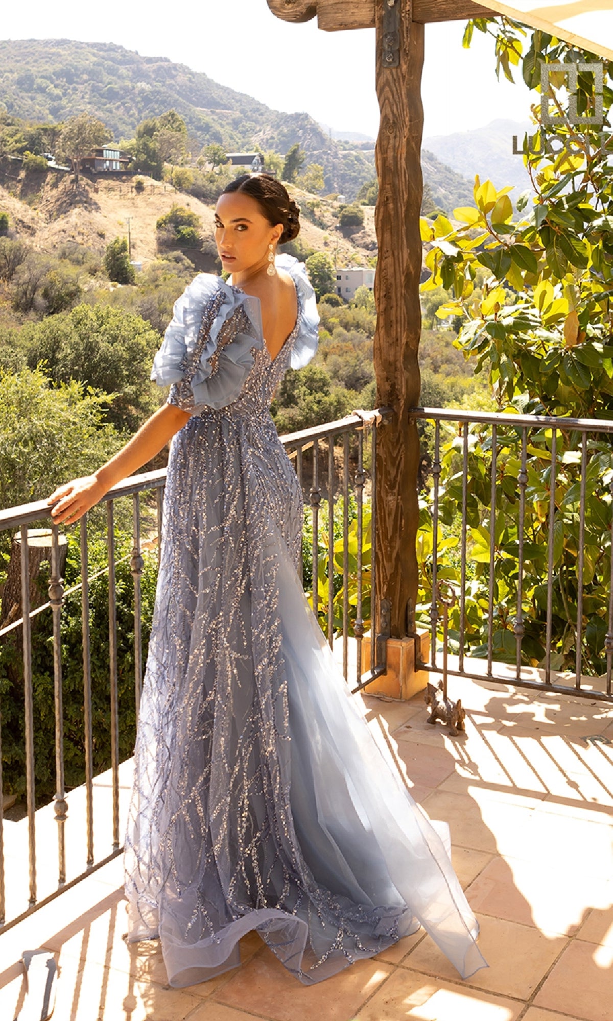 Wearing the Lucci Lu C8042 Long Prom Dress in blue, a woman with ruffled sleeves and a semi-sheer bodice stands on a sunlit balcony overlooking scenic hills.
