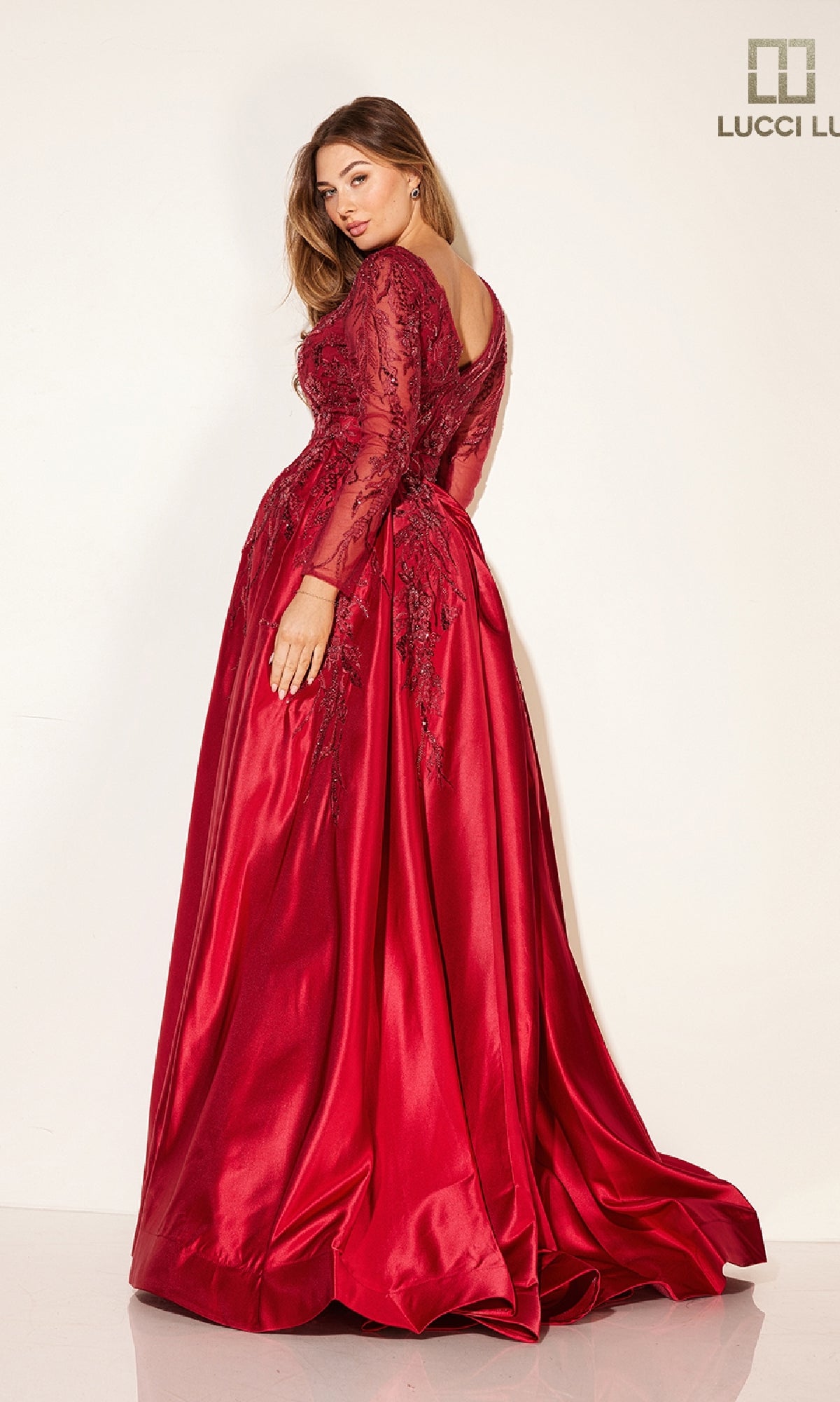 A woman models the Long Formal Dress: Lucci Lu C8045, a red satin gown with sparkly embroidery, lace detailing, and long sleeves, while posing against a plain light background.
