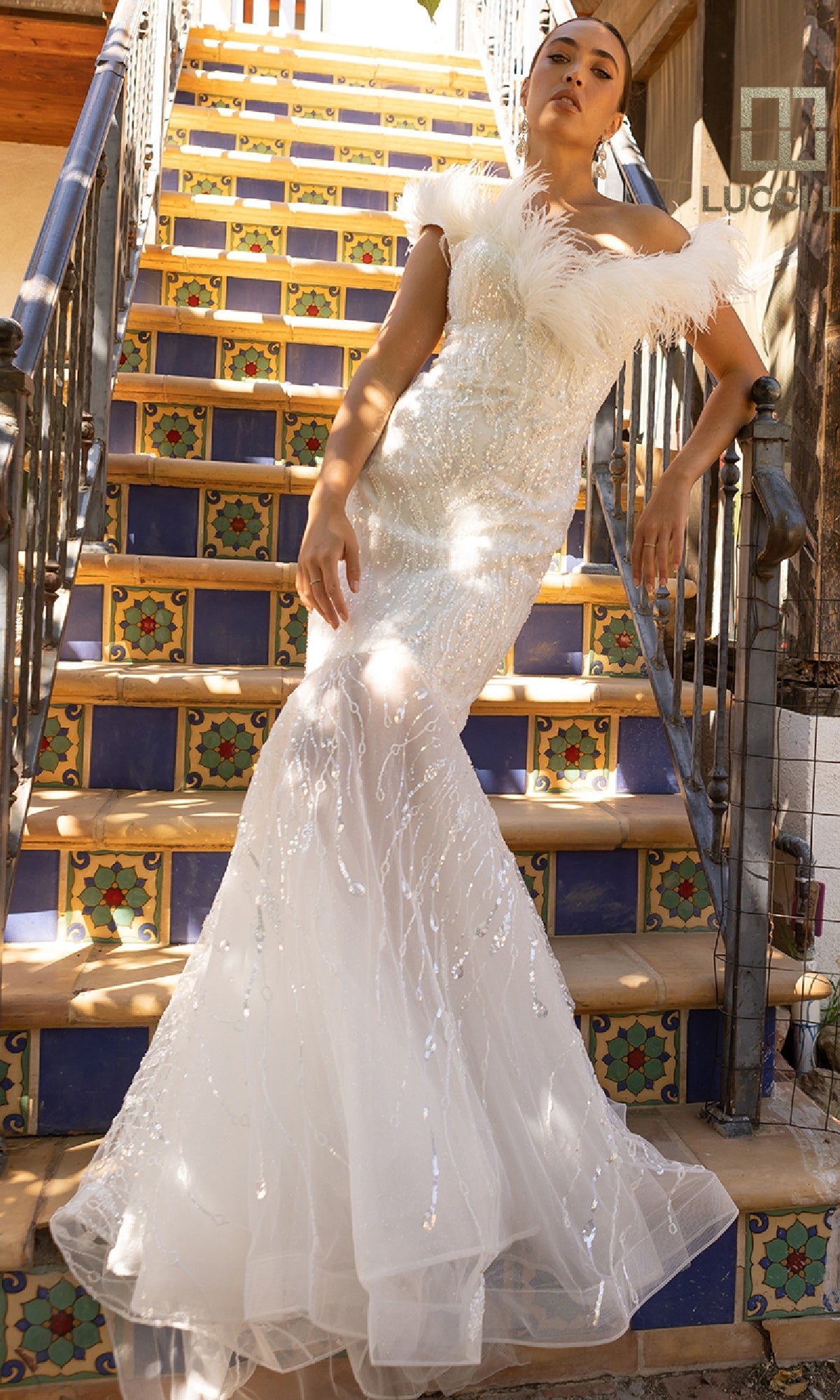 Wearing the Beaded Tulle White Wedding Dress Lucci Lu C8051, a woman poses on decorative tiled stairs with one hand on the railing.