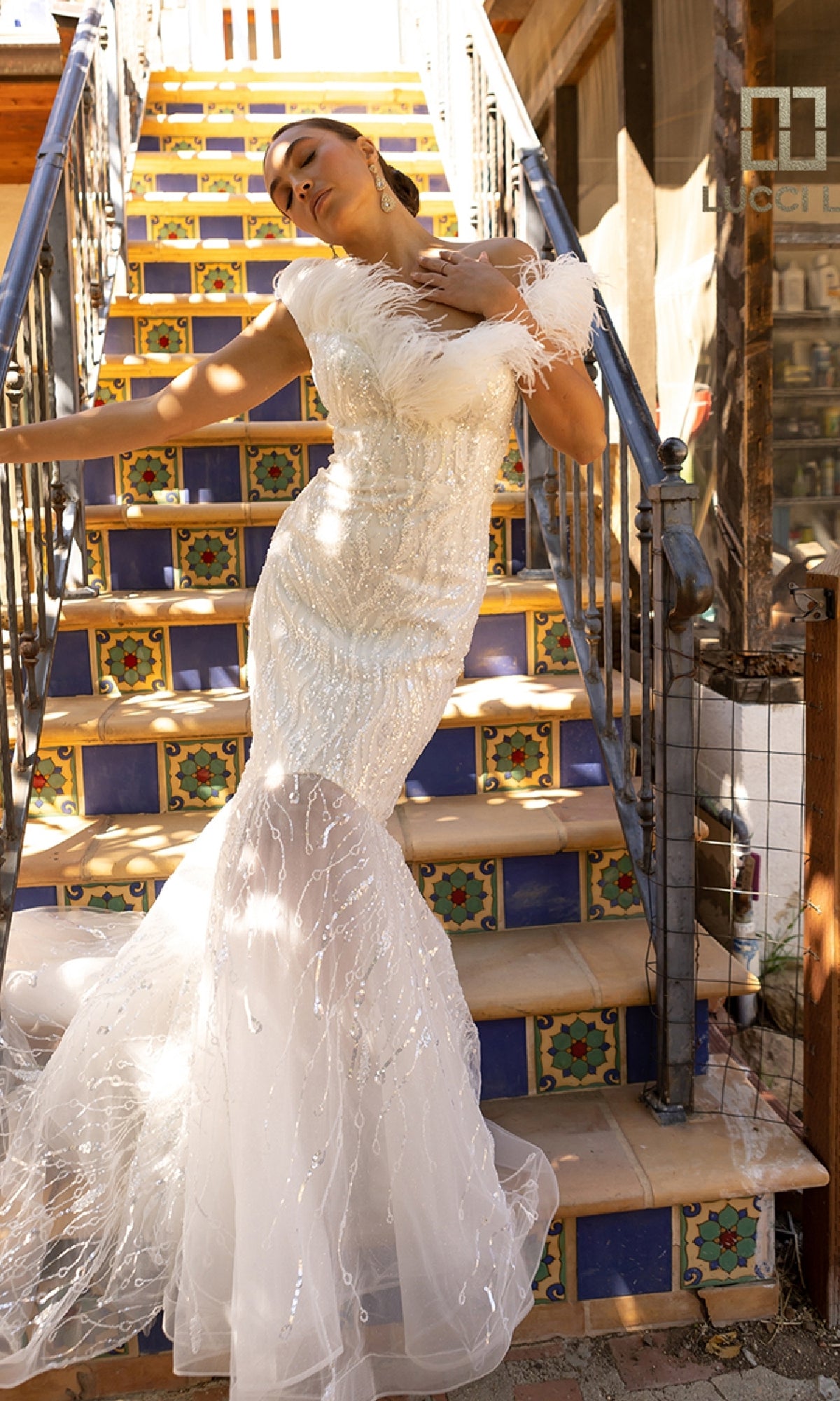 A woman wearing the Beaded Tulle White Wedding Dress Lucci Lu C8051 poses on colorful tiled stairs, one hand on her chest and her head tilted back.