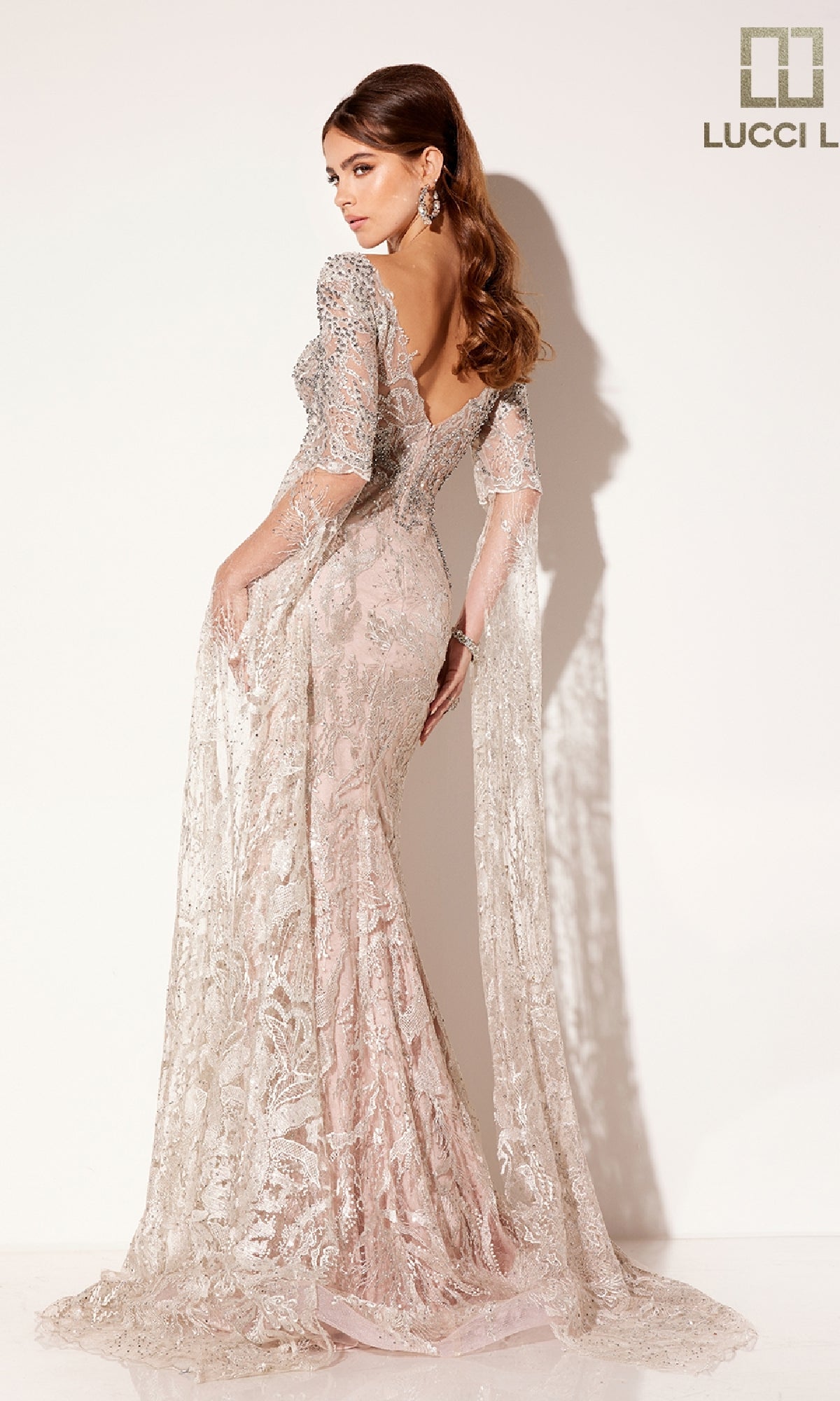 A woman models the Lucci Lu C8073 long formal dress—a fitted, floor-length pastel pink lace gown with sheer, embellished sleeves and a low back—against a plain background featuring the