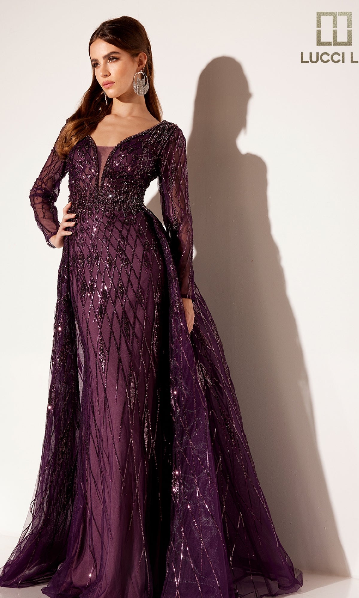 A woman stands elegantly against a light background wearing the Lucci Lu C8095, a long formal dress in deep purple with sequin detailing and a sheer tulle overskirt.