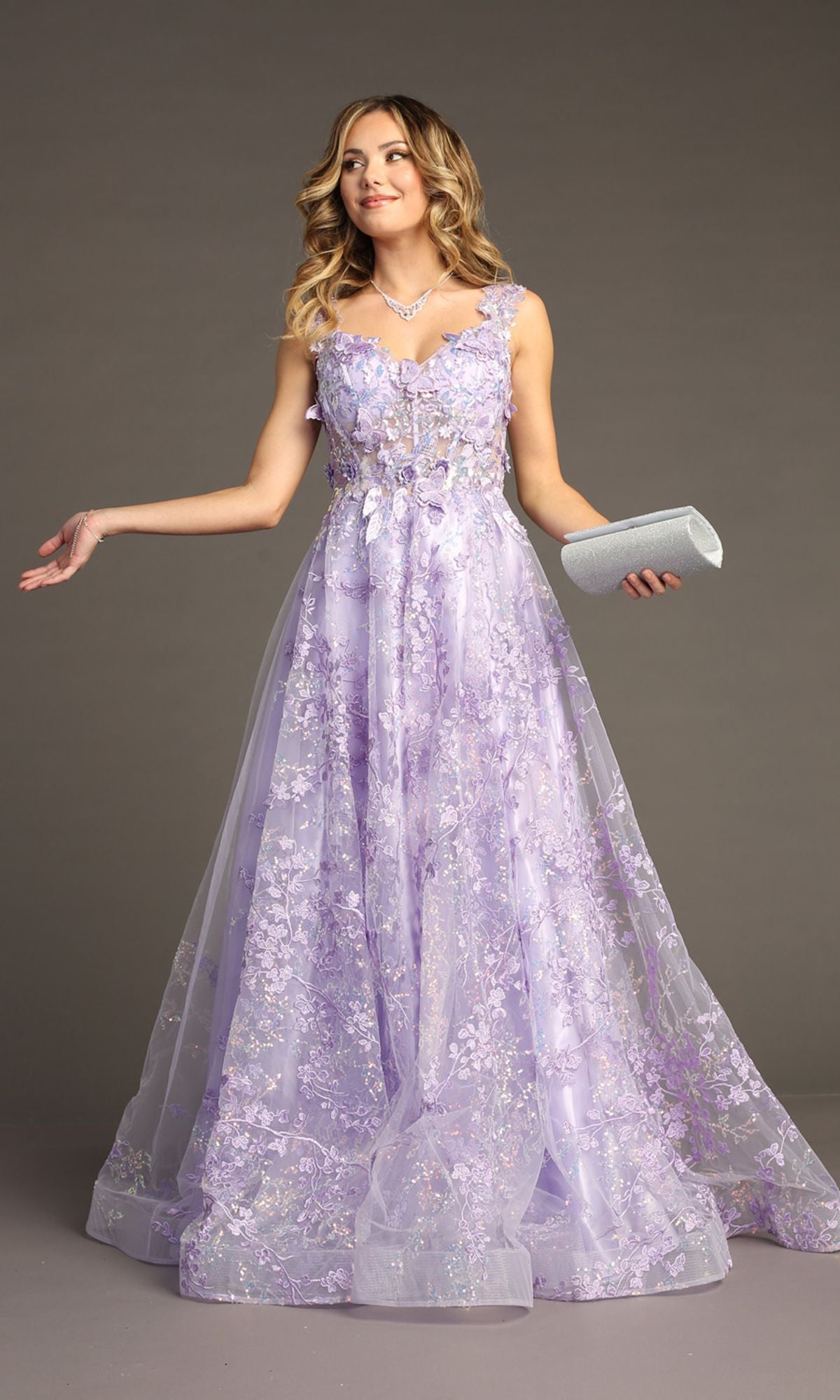 A woman wears the Floral-Print Lace-Up Long Prom Dress: Chicas C818, a floor-length light purple gown with a corset-style bodice, holding a silver clutch against a plain gray background.