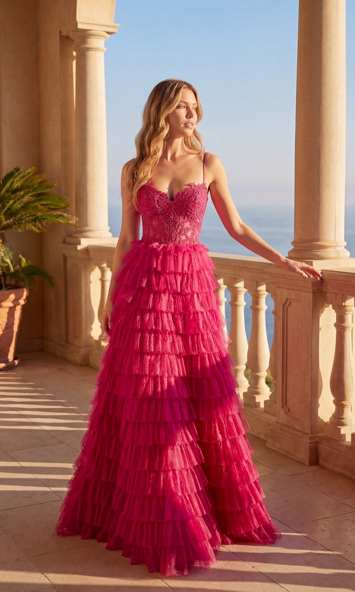 A woman in the fuchsia Long Prom Dress: Chicas C833 with tiered ruffles, spaghetti straps, and a lace-up back stands on a stone balcony overlooking the ocean.