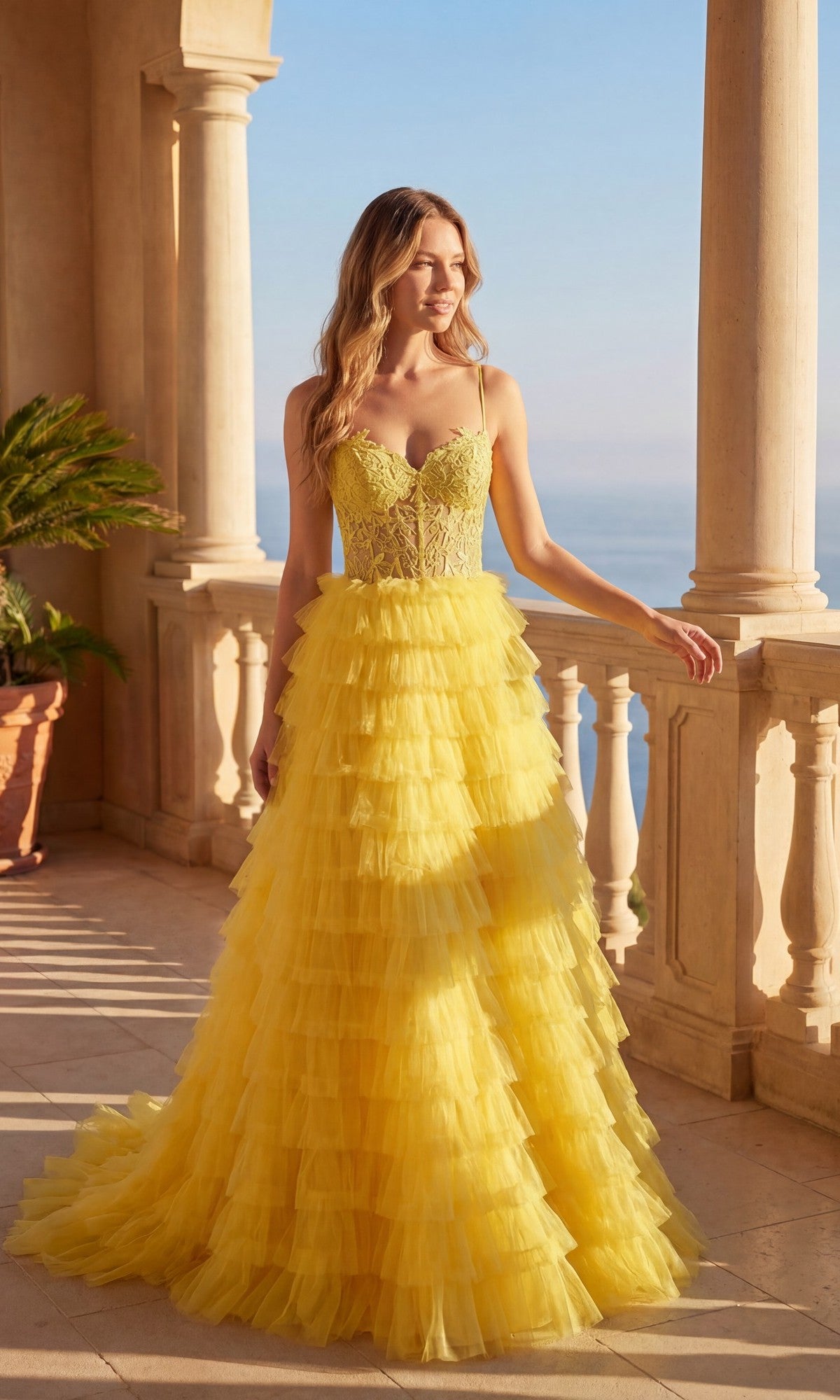 A woman stands on a stone balcony in daylight, wearing the Chicas C833 Long Prom Dress—a yellow tiered ruffle ball gown with a lace embroidered bodice and lace-up back—against an ocean and clear sky backdrop.
