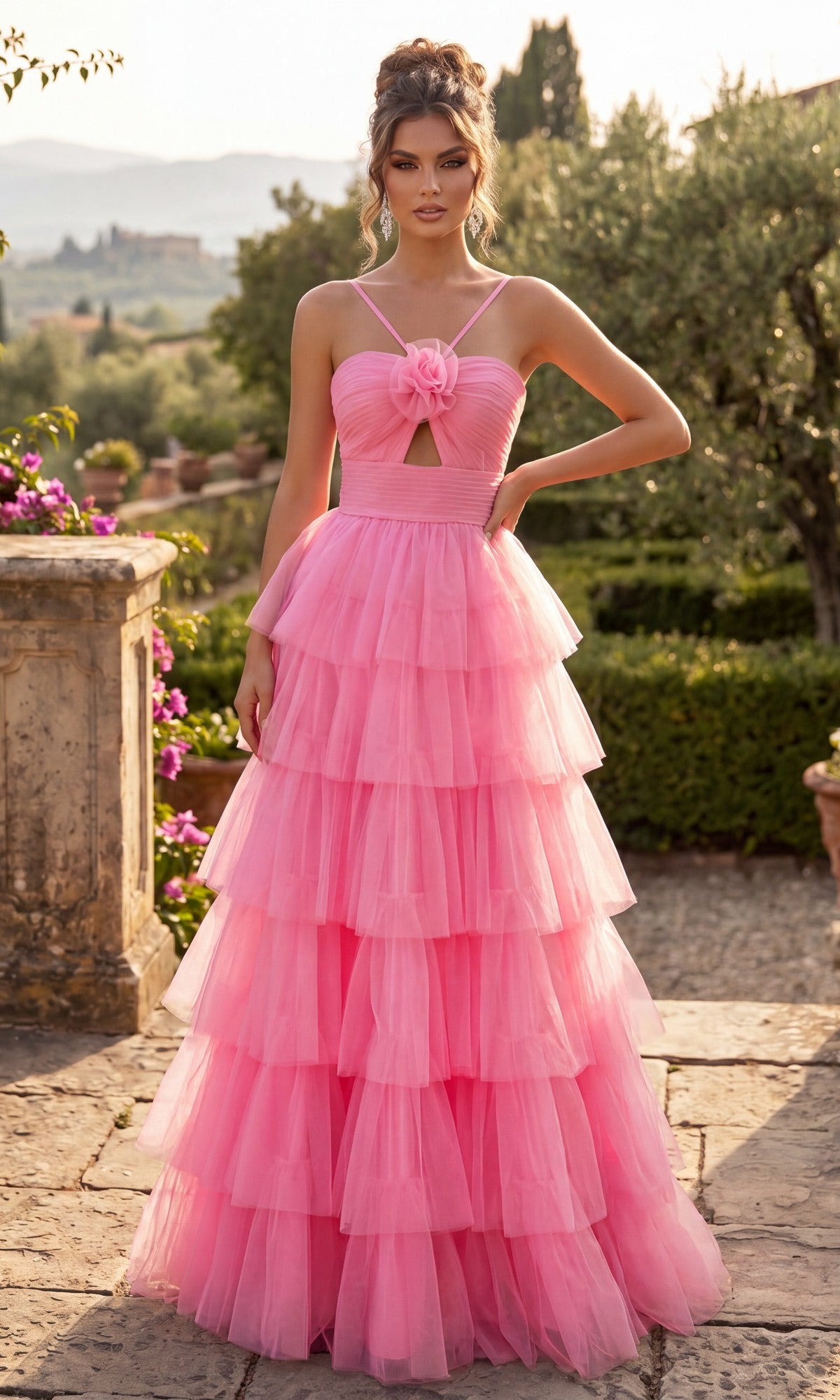 A woman stands outdoors amid lush greenery and distant buildings, wearing the Chicas C837 Long Prom Dress in pink—a floor-length, sleeveless ball gown crafted from layered tulle with a floral detail on the bodice and a dramatic, tiered full skirt.