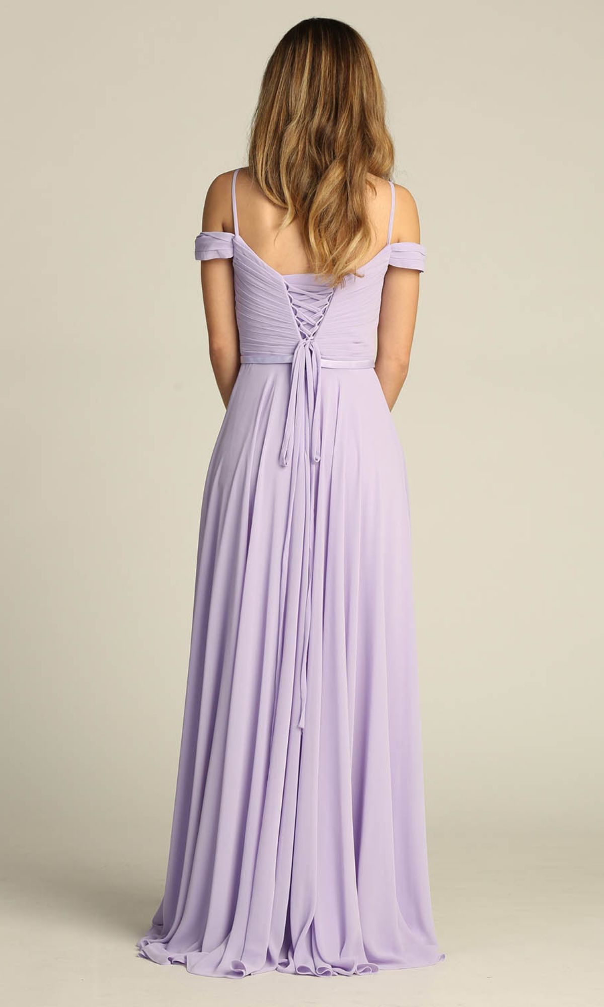 A woman with long wavy hair models the Chicas C8997 Long A-Line Off-Shoulder Prom Dress in lavender, featuring a lace-up back and floor-length skirt, standing with her back to the camera.