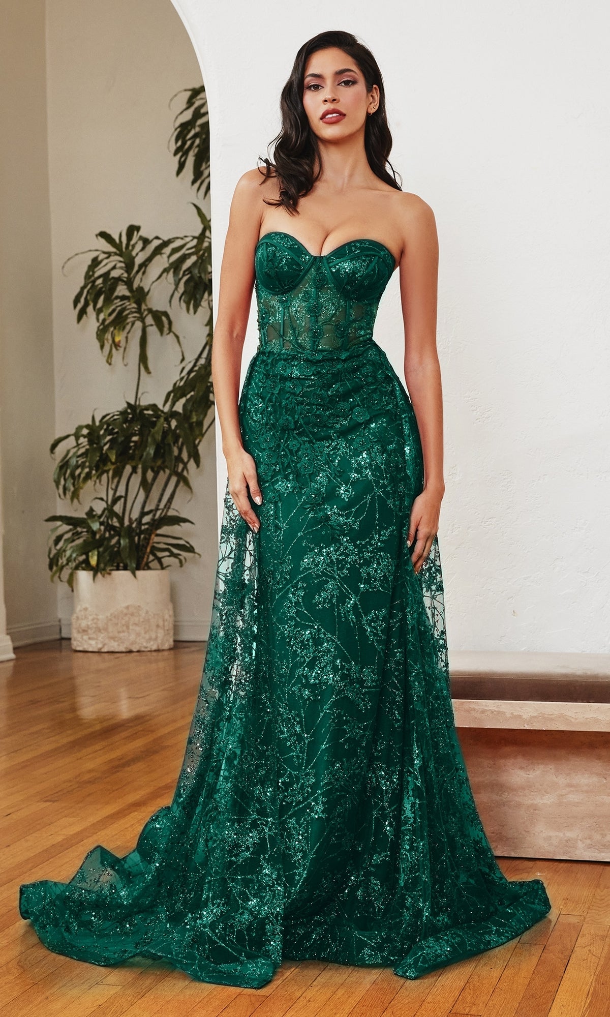A woman stands indoors wearing the Glitter-Print Strapless Corset Prom Dress CB046 in emerald green, showcasing a sheer bodice with intricate glitter-print details and floor-length elegance.